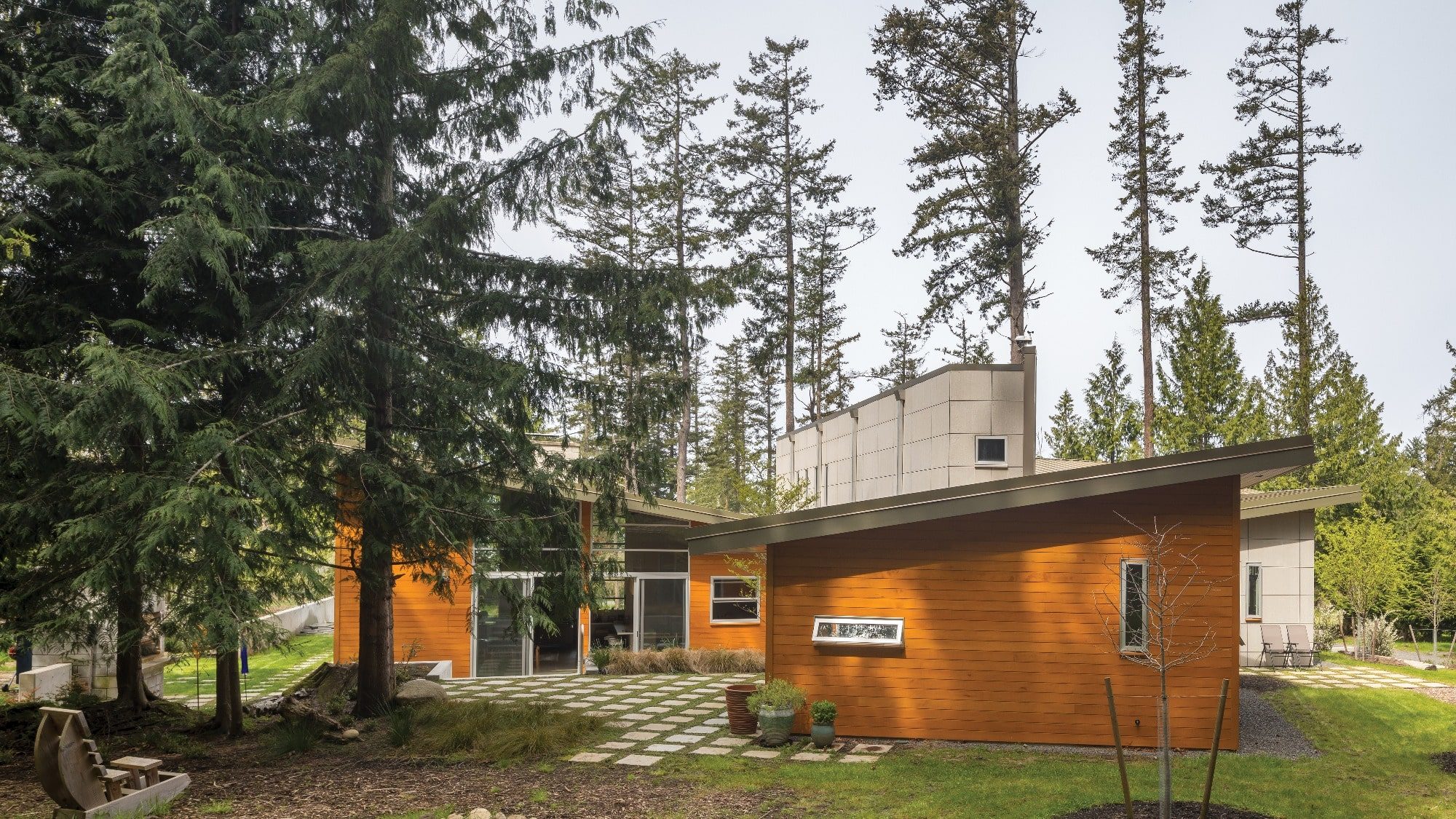 Modern house with wood siding and large windows surrounded by tall trees, embodying the Seattle home design aesthetic. A stone pathway and small garden are in the foreground, adding charm to the exterior that complements an interior design focused on natural light.
