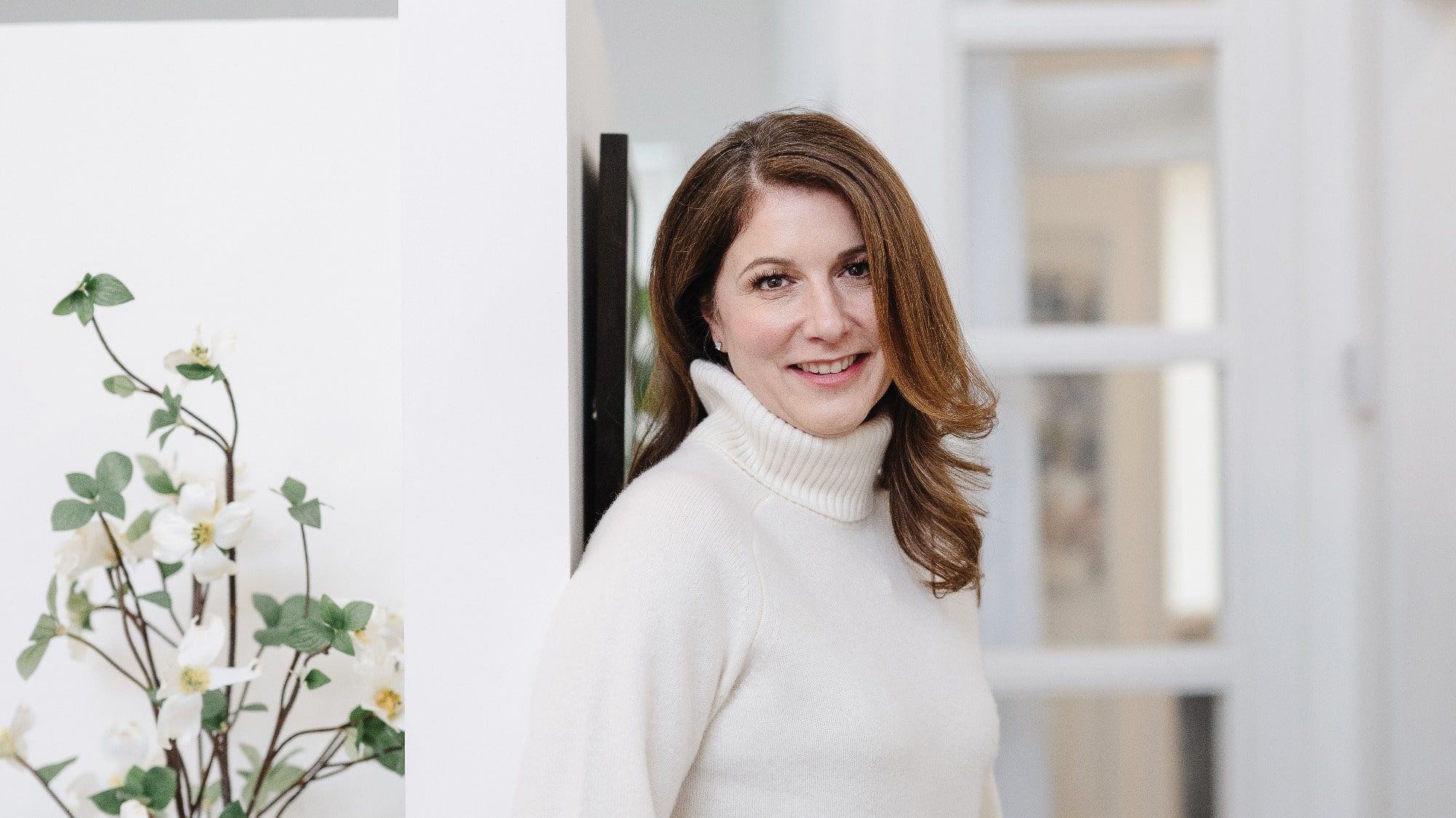 A woman in a white turtleneck sweater leans against a wall near a doorframe, embodying the Seattle home design aesthetic. A plant with white flowers graces the foreground, adding a touch of nature to this elegantly curated interior design space.