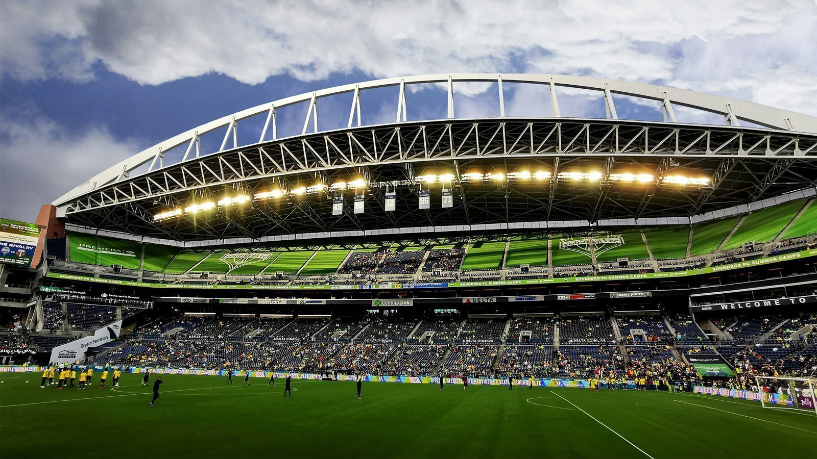 Wide-angle view of a large, illuminated stadium with a curved roof and green seating, buzzing with the energy of Seattle sports fans. A crowd gathers as a group stands on the green field, surrounded by digital screens displaying vibrant imagery.