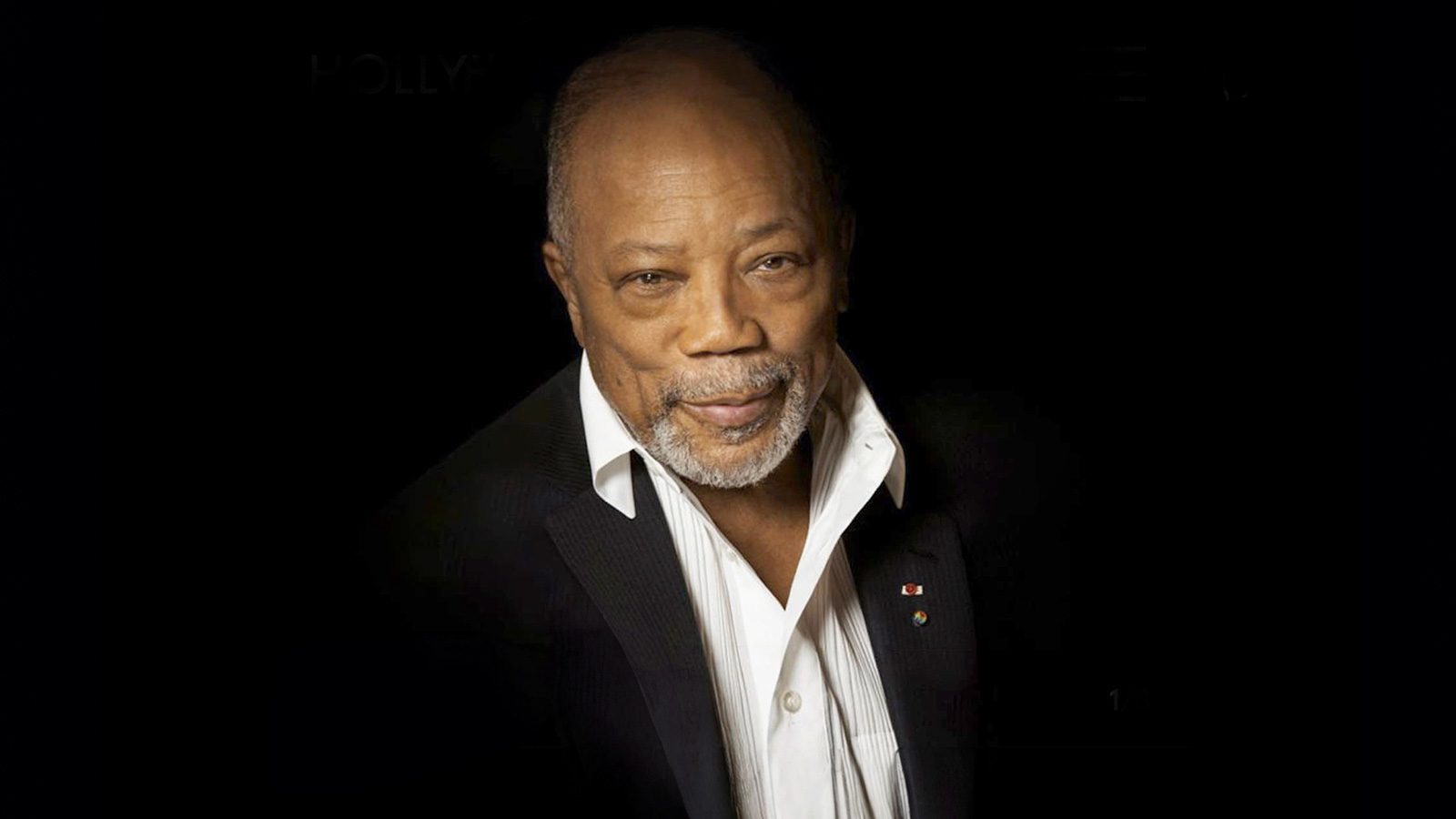 A man with a beard and a mustache, wearing a white shirt and dark jacket, smiles against a dark background.