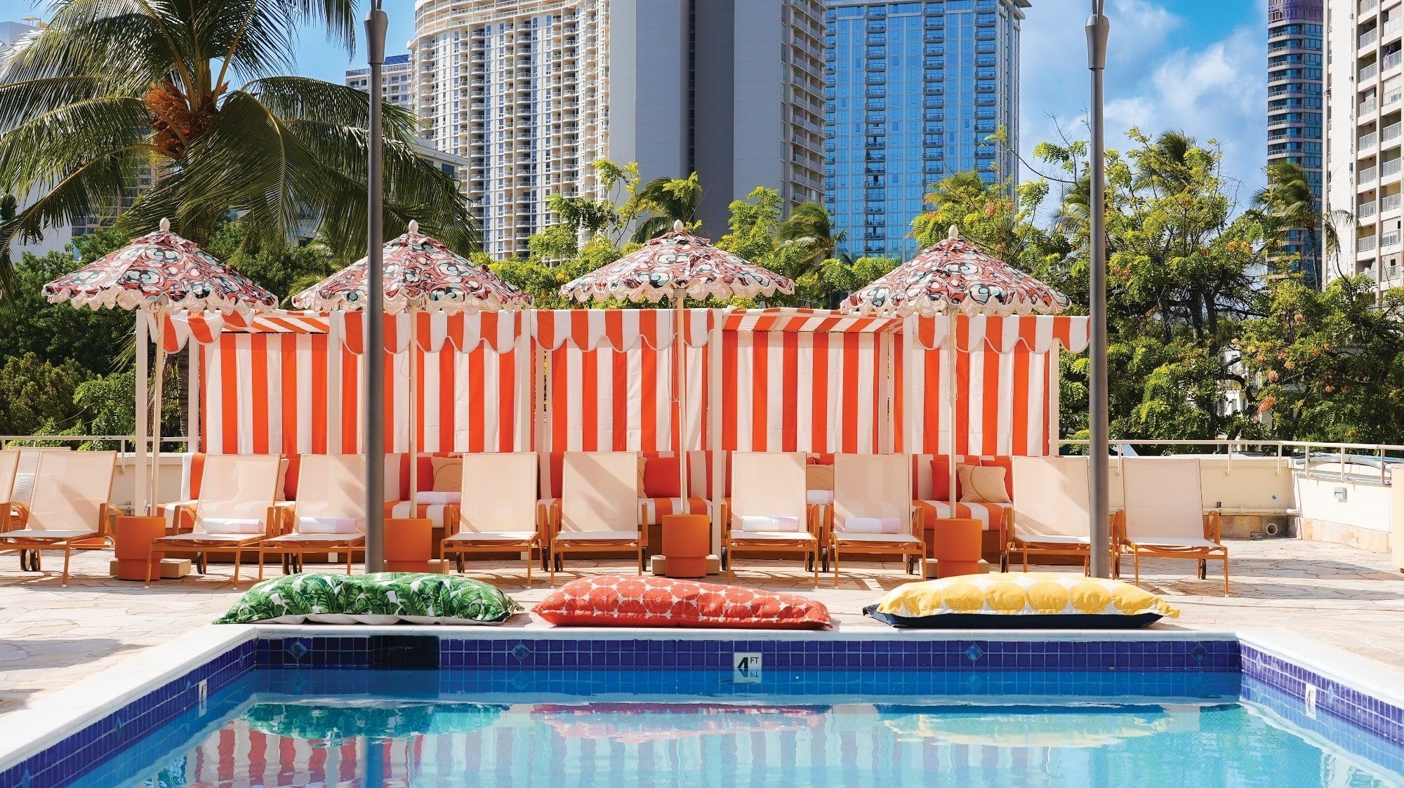 The poolside area in Waikiki boasts striped umbrellas, lounge chairs, and colorful cushions. Tall city buildings and trees create a scenic backdrop under the light of a clear blue sky.
