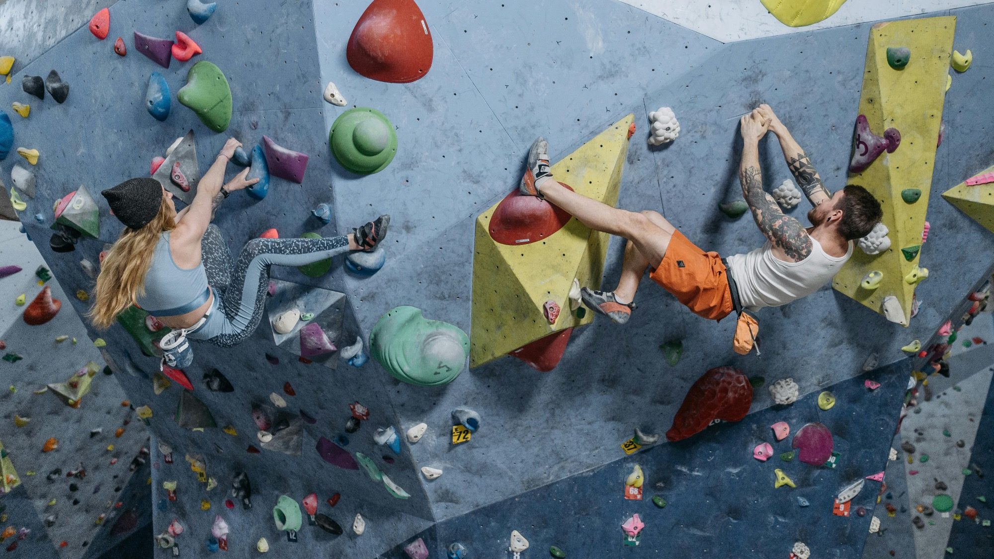 Amidst the Games of Gravity, two individuals tackle the indoor climbing wall, navigating colorful holds in their athletic gear.