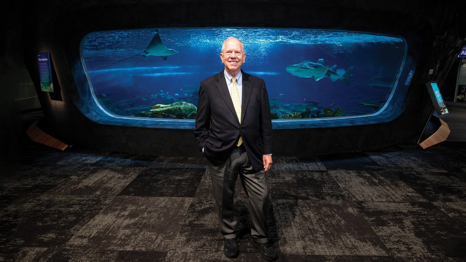 Bob Davidson visited the Seattle Aquarium, where a person in a suit stands in front of a large tank brimming with diverse fish and marine animals.