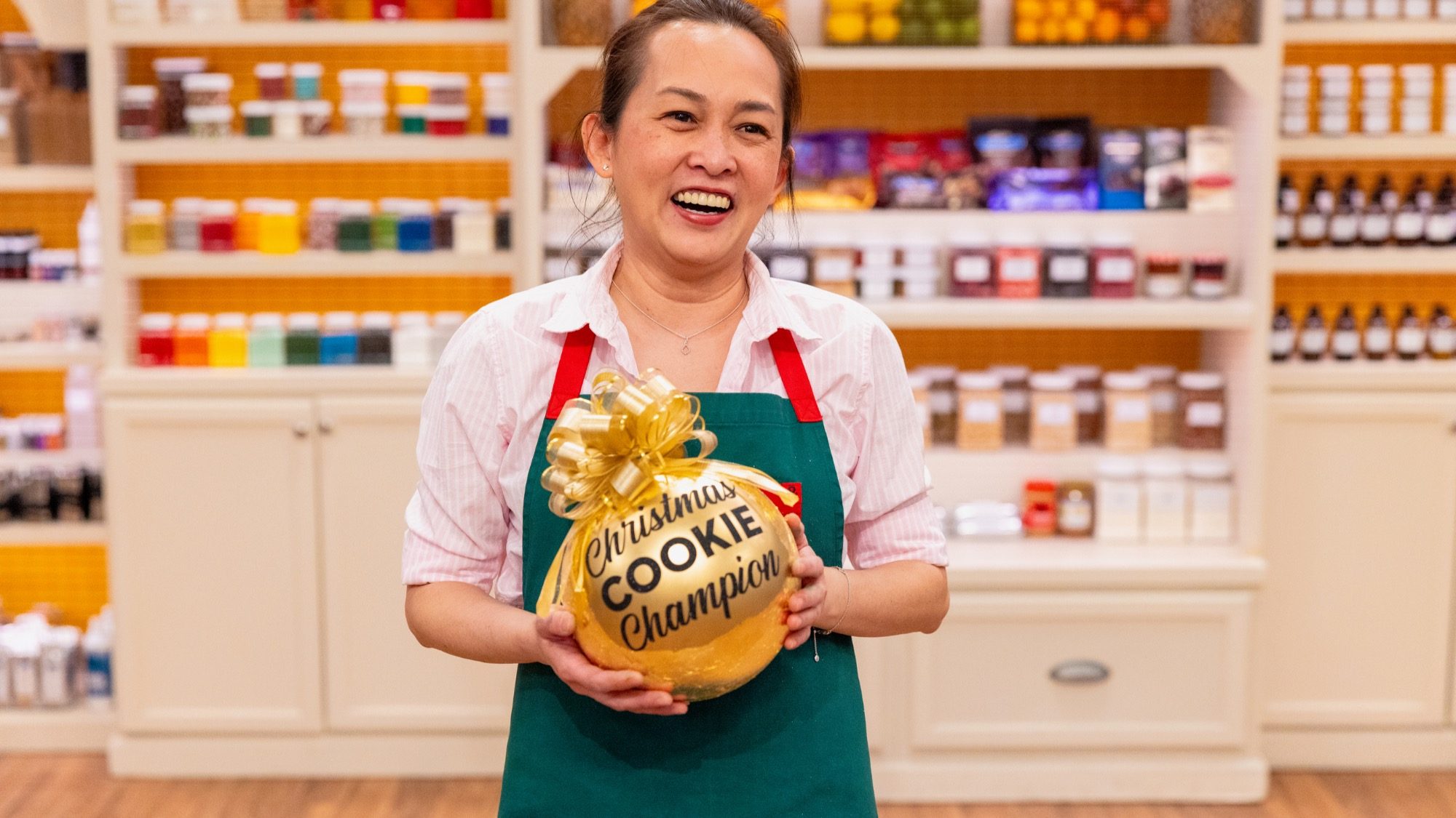 A Seattle baker wearing an apron proudly holds a golden ornament inscribed with "Christmas Cookie Champion" after excelling in the Food Network's Cookie Challenge, standing amidst shelves brimming with ingredients in their kitchen.