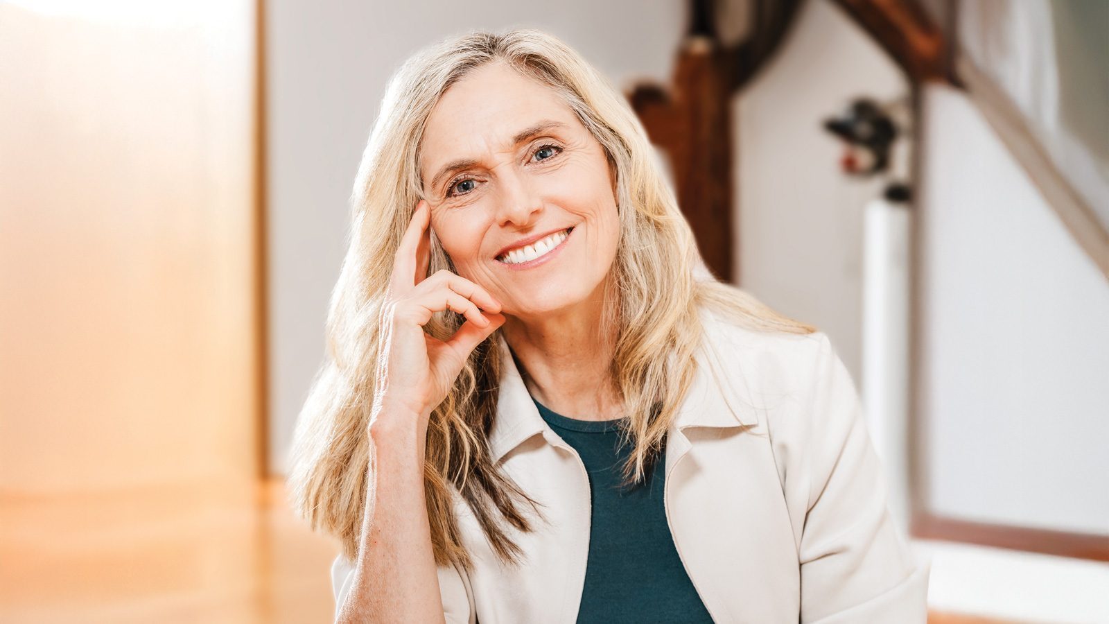 Delaney Ruston, a woman with long blond hair, smiles gently while resting her head on her hand. She wears a light jacket over a dark top, exuding warmth and charm.