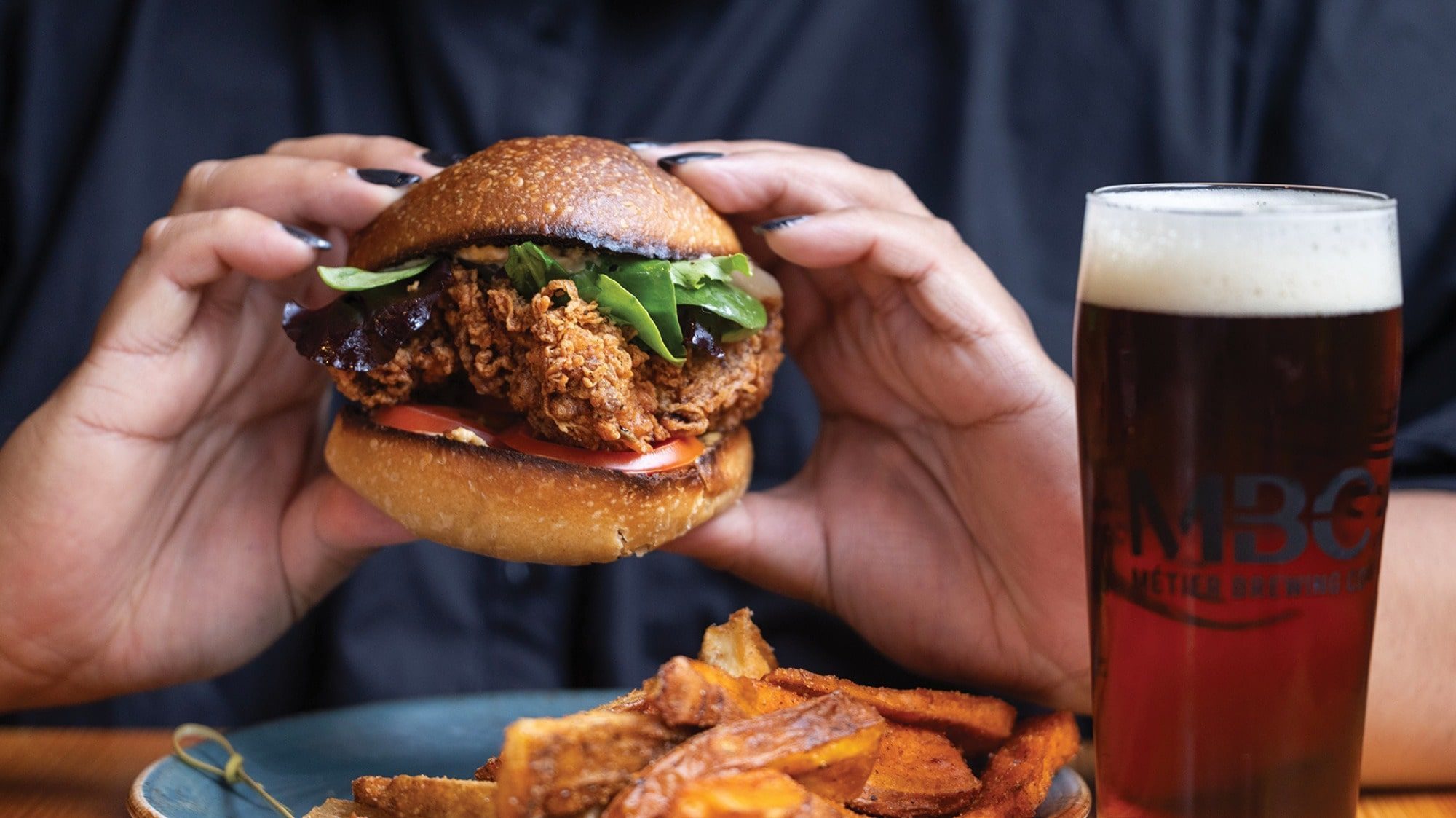 Person holding a fried chicken sandwich with lettuce and tomato at Seattle's hot attraction. A plate of fries and a glass of dark beer sit nearby, creating the perfect meal before heading out on a unique hot tub boat adventure.
