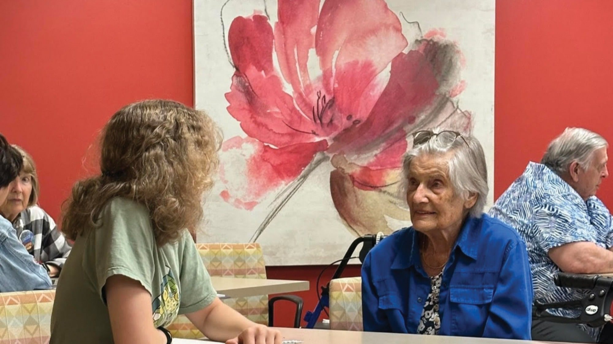 A young person and an older woman sit across from each other at a table in a room with red walls and a large floral painting, their conversation filled with stories and aspirations as kids look toward the future.