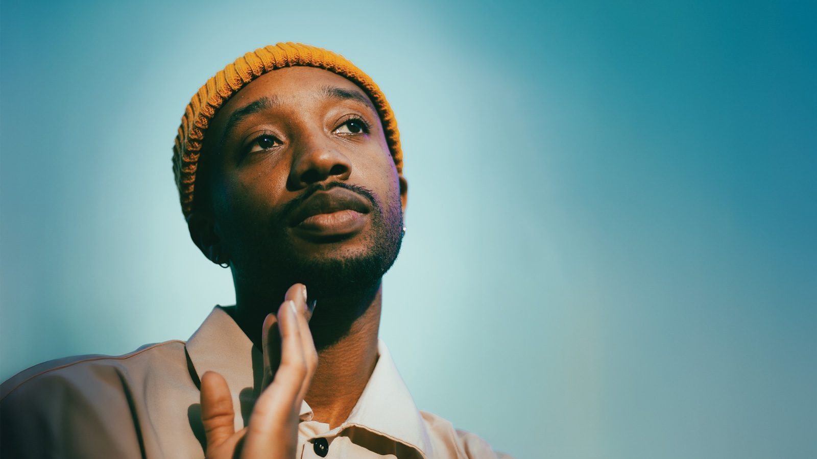 Person wearing a yellow beanie and light-colored shirt, looking upward with a thoughtful expression against a gradient blue background, reminiscent of the introspective gaze often associated with Poet Luther Hughes.