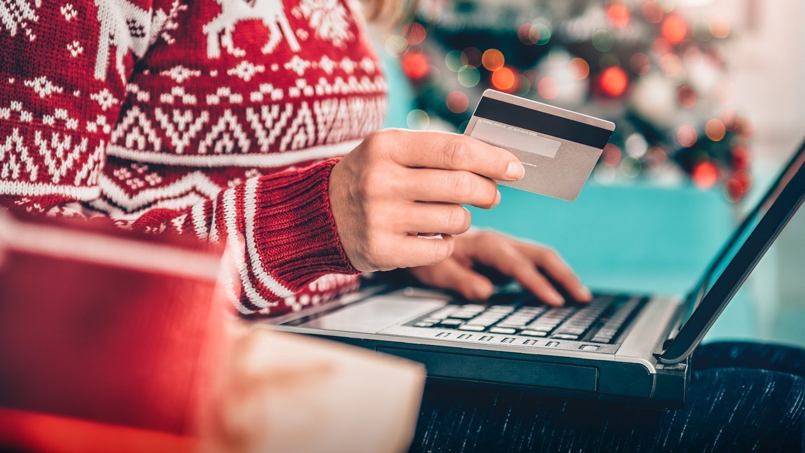 A cheerful individual in a festive sweater holds a credit card, eagerly browsing Seattle mag’s last-minute holiday travel gift guide on their laptop, with the glow of a blurred Christmas tree painting a cozy backdrop.