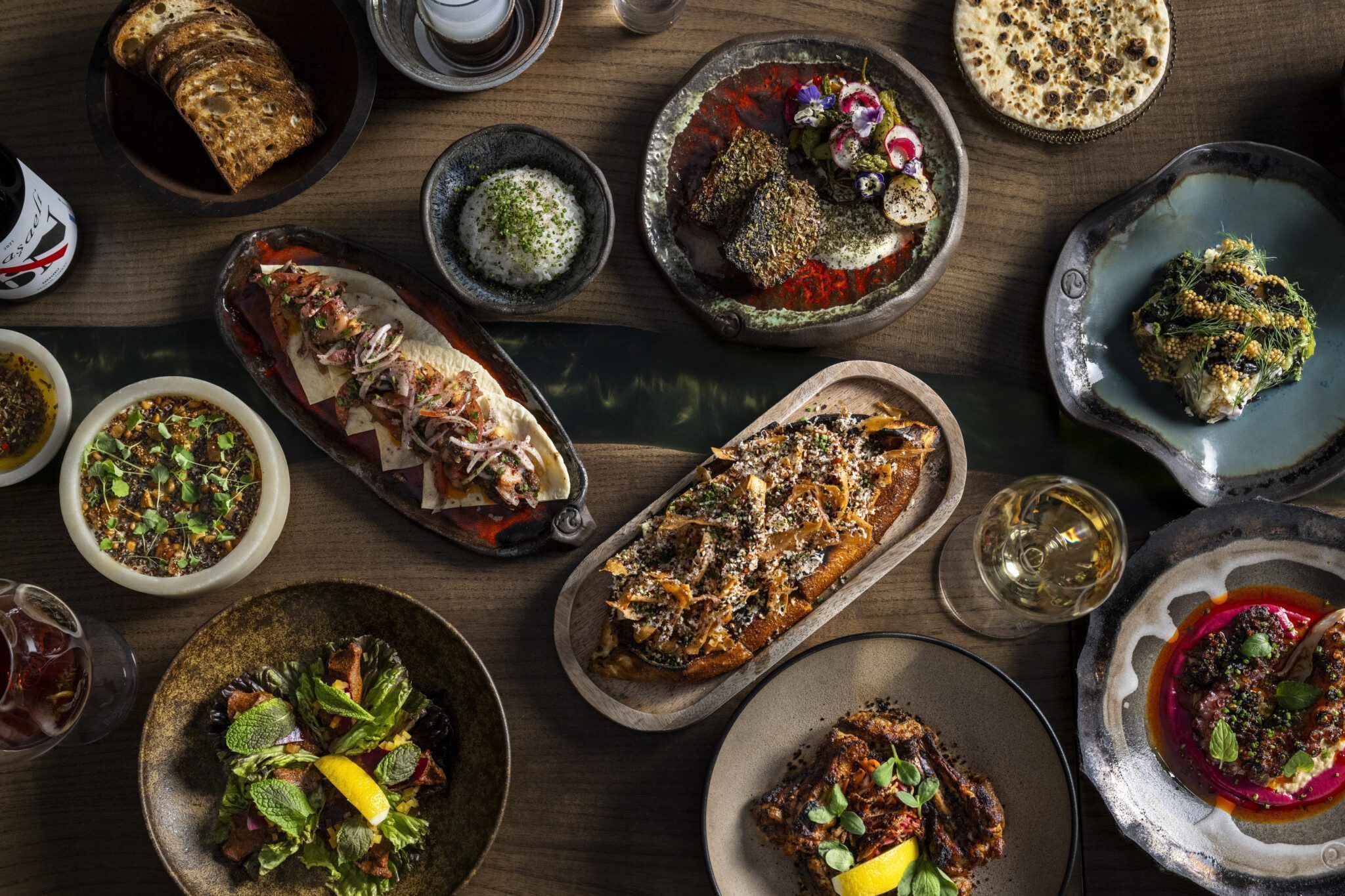 A table with various dishes, including grilled meats, vegetables, salads, dips, and bread, alongside glasses of wine and water.