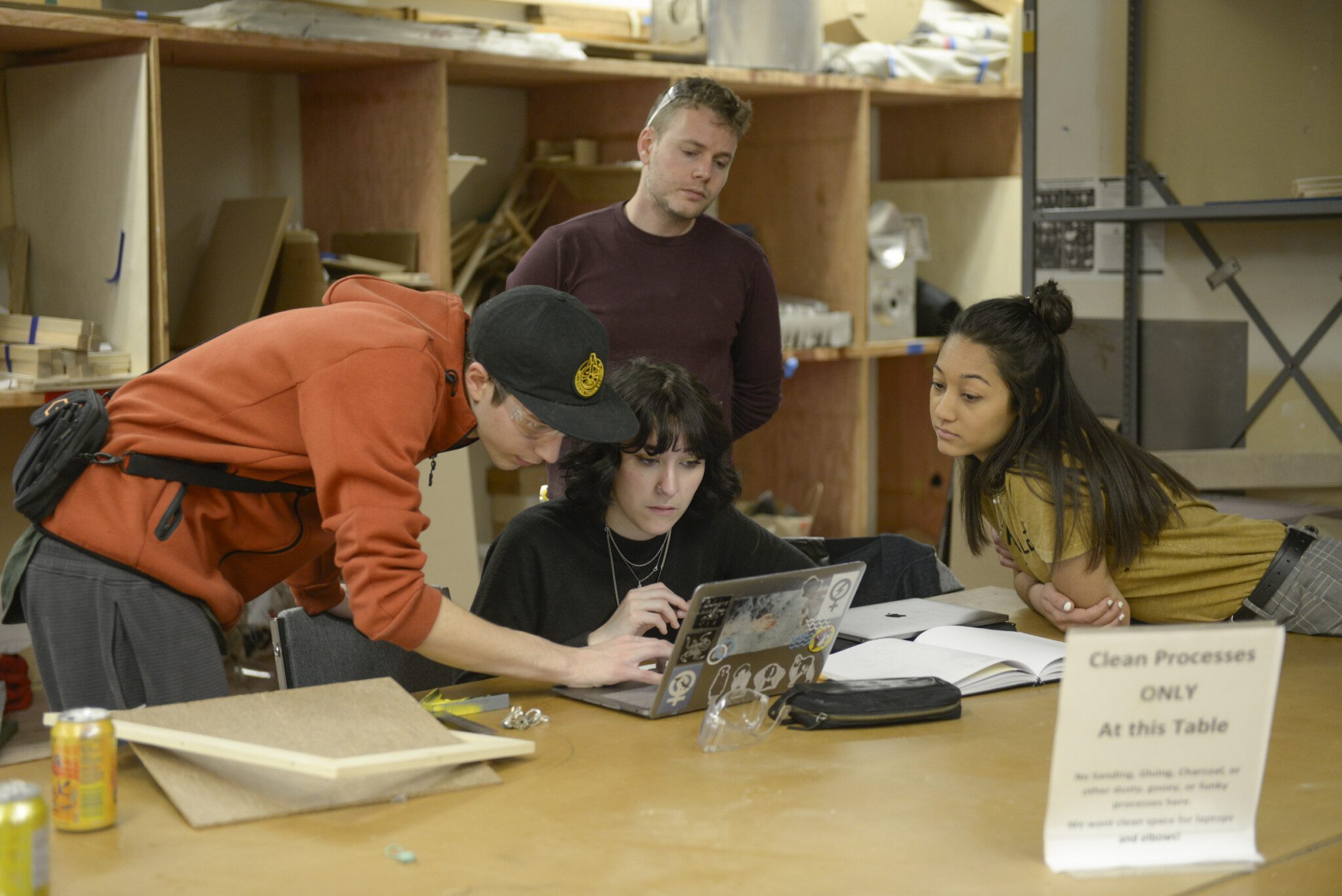 Four people in a workshop setting are gathered around a laptop on a table with tools and materials. A sign reads "Clean Processes ONLY At this Table.