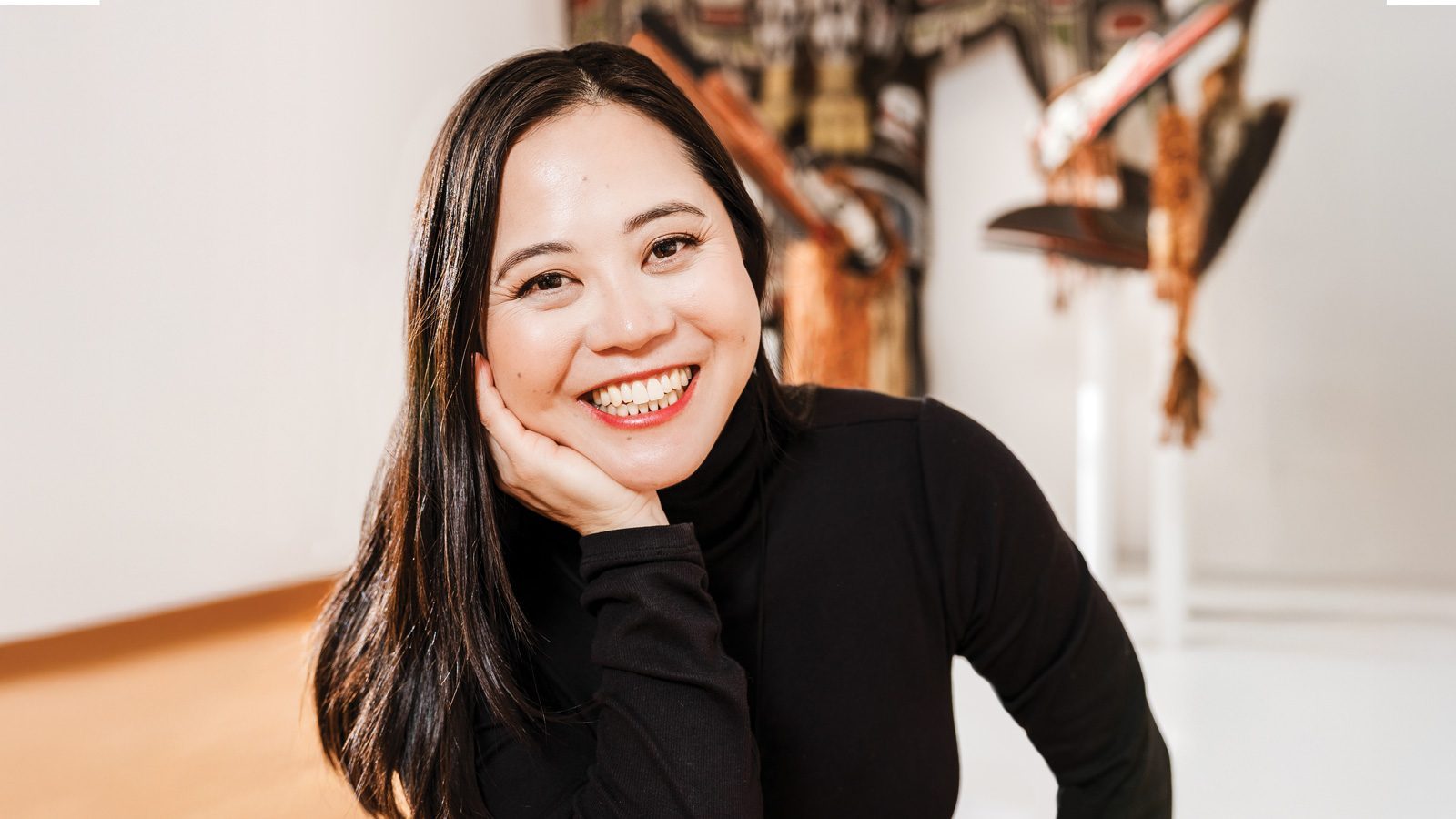 A Vietnamese American author, Susan Lieu, smiles gently while resting her chin on her hand, her long black hair cascading over a black long-sleeve top.