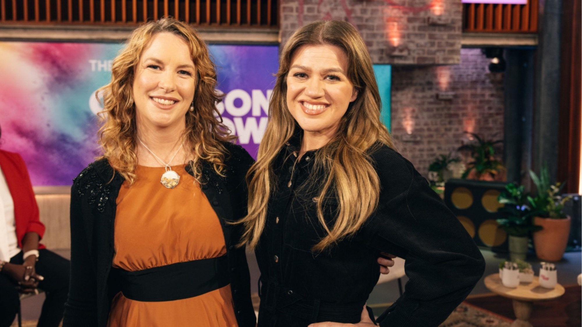 Two women, one in an orange dress and the other in a black top, smile and pose indoors against a colorful backdrop with a brick wall. Their joyful vibes could light up any room. It's like they've just found out they're the next potential winners of $100,000 for supporting Pike Place Market Foundation!