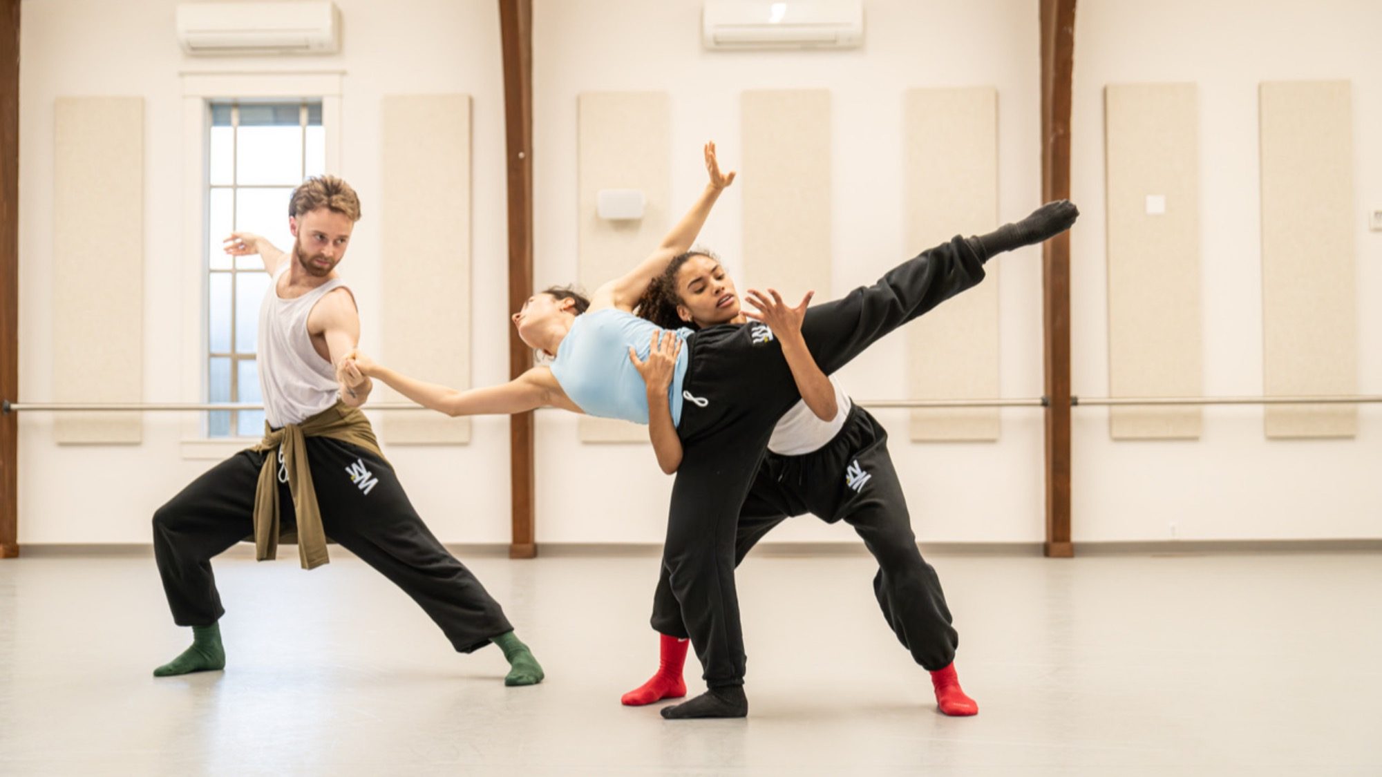 Three dancers perform a contemporary routine in a studio, each in dynamic poses. The two in black pants support the dancer in blue, who is extending their leg high.