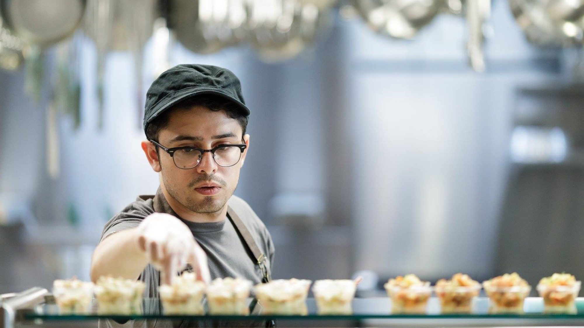 A person wearing glasses and a cap arranges small cups of food on a counter in a kitchen.