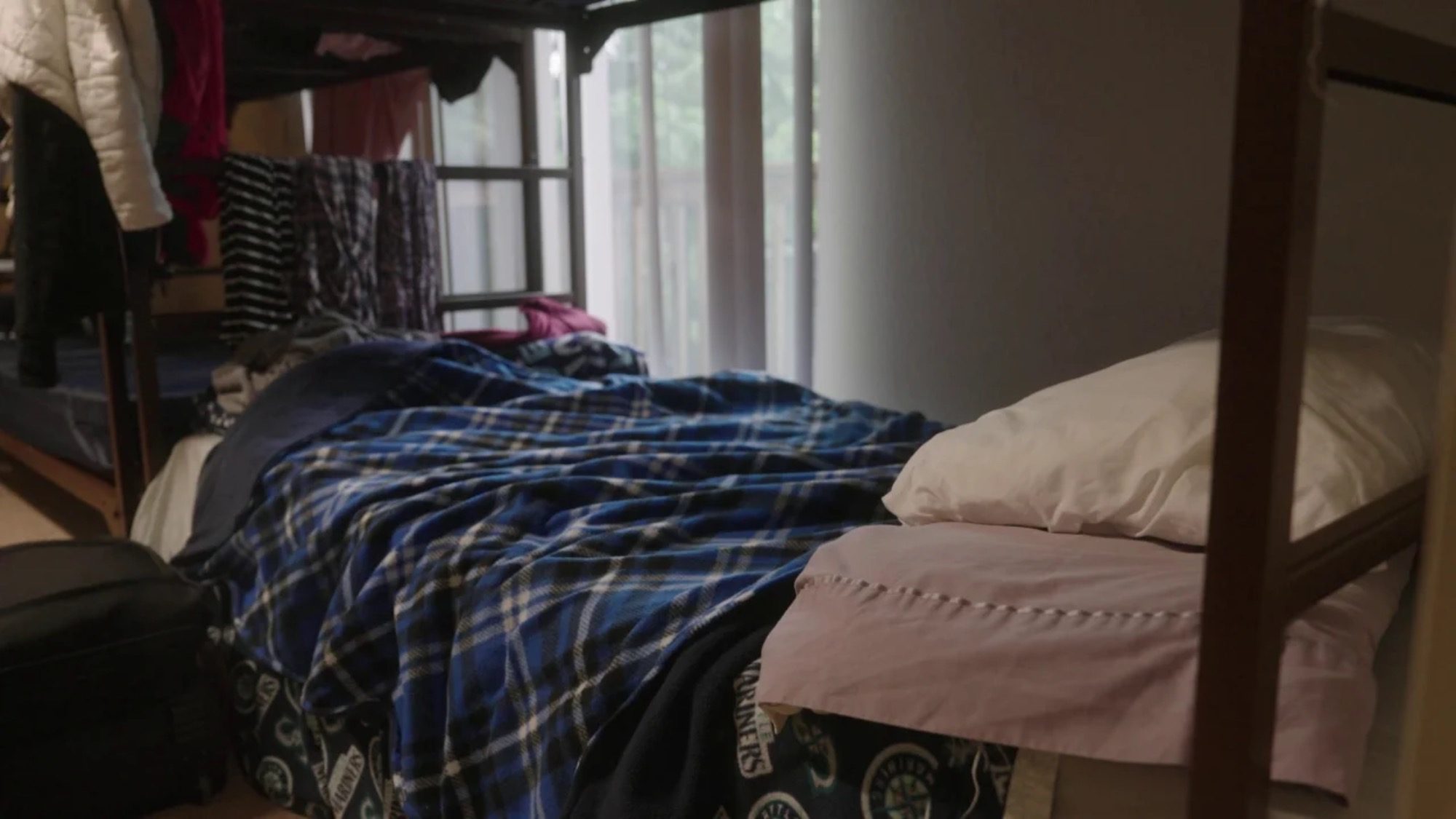 A bunk bed adorned with a blue plaid blanket and a white pillow sits beside a window with sheer curtains, reminding us that every step counts in addressing the issues that persist.