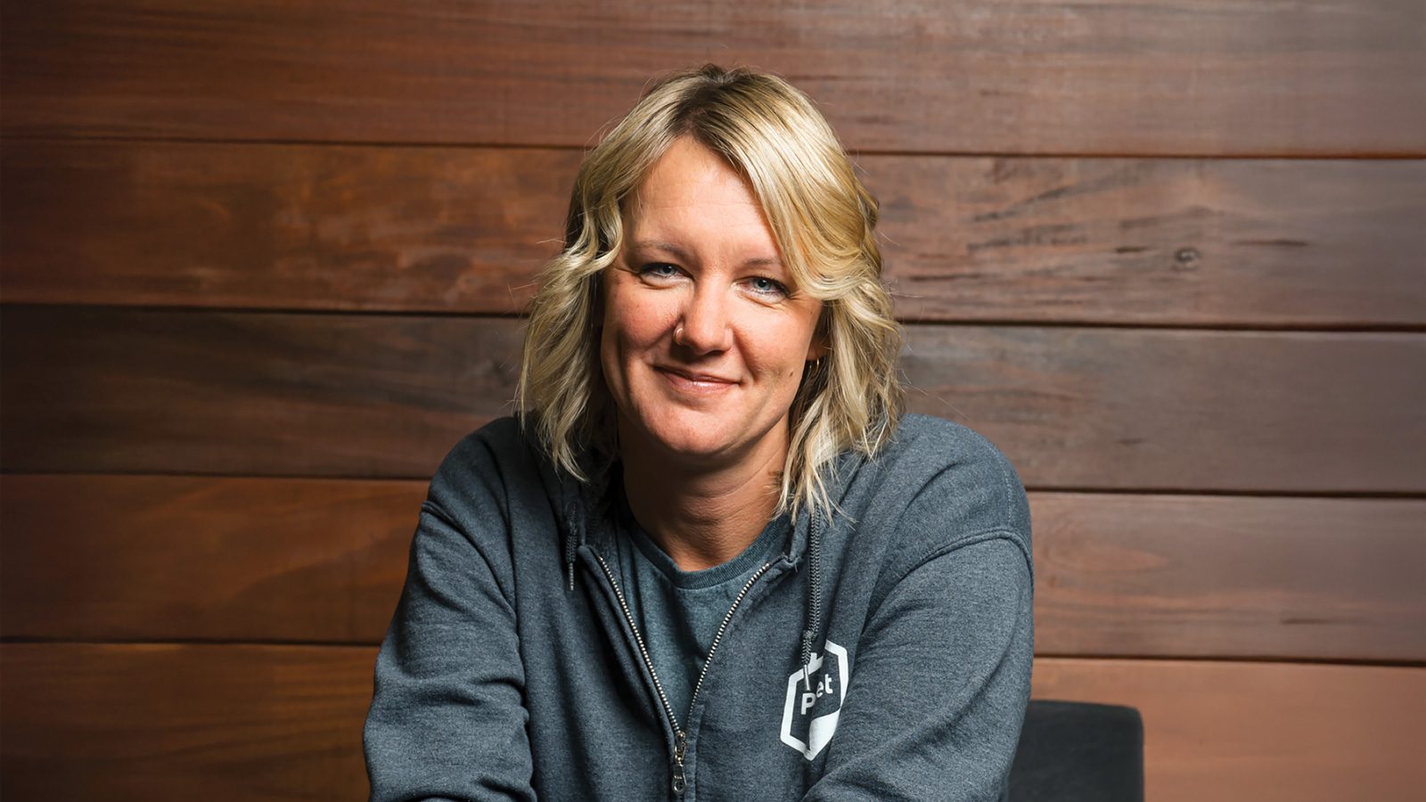 A person with blonde hair in a gray hoodie smiles in front of a wooden background.