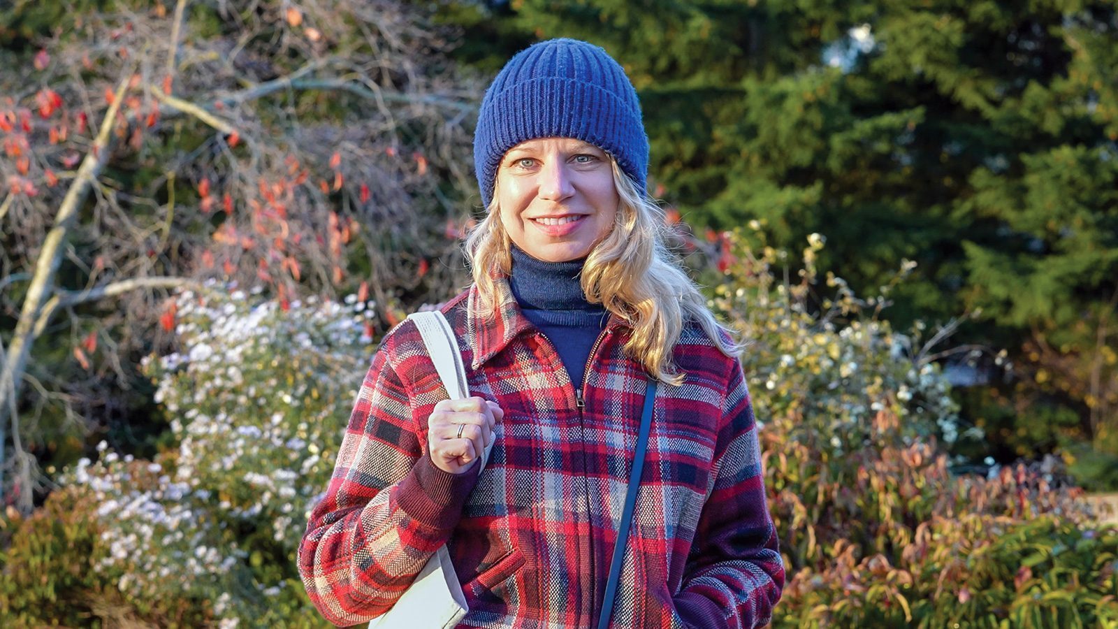A person in a blue hat and red plaid jacket stands outdoors, holding a white bag. Trees and autumn foliage are in the background.