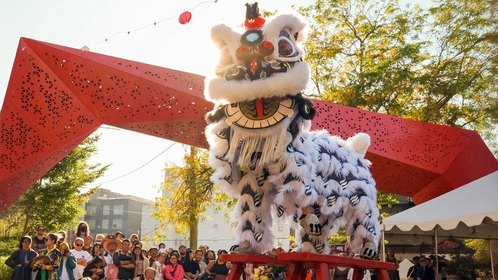 A traditional lion dance performance, featuring a white lion costume, takes place under a red geometric archway adorned with vibrant red lanterns, as a crowd of onlookers gathers beneath the gentle rain.