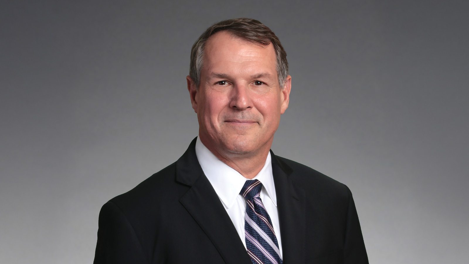 Dave Bateman, dressed sharply in a black suit and striped tie, poses confidently against a gray background.