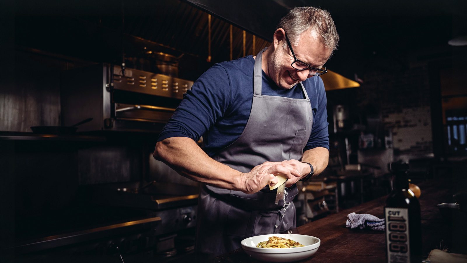 In a bustling kitchen, a person wearing glasses and an apron grates cheese over a bowl of pasta, surrounded by gleaming kitchen equipment. It's a scene where the belief that good food can change lives comes to life with every delicious twist of the grater.