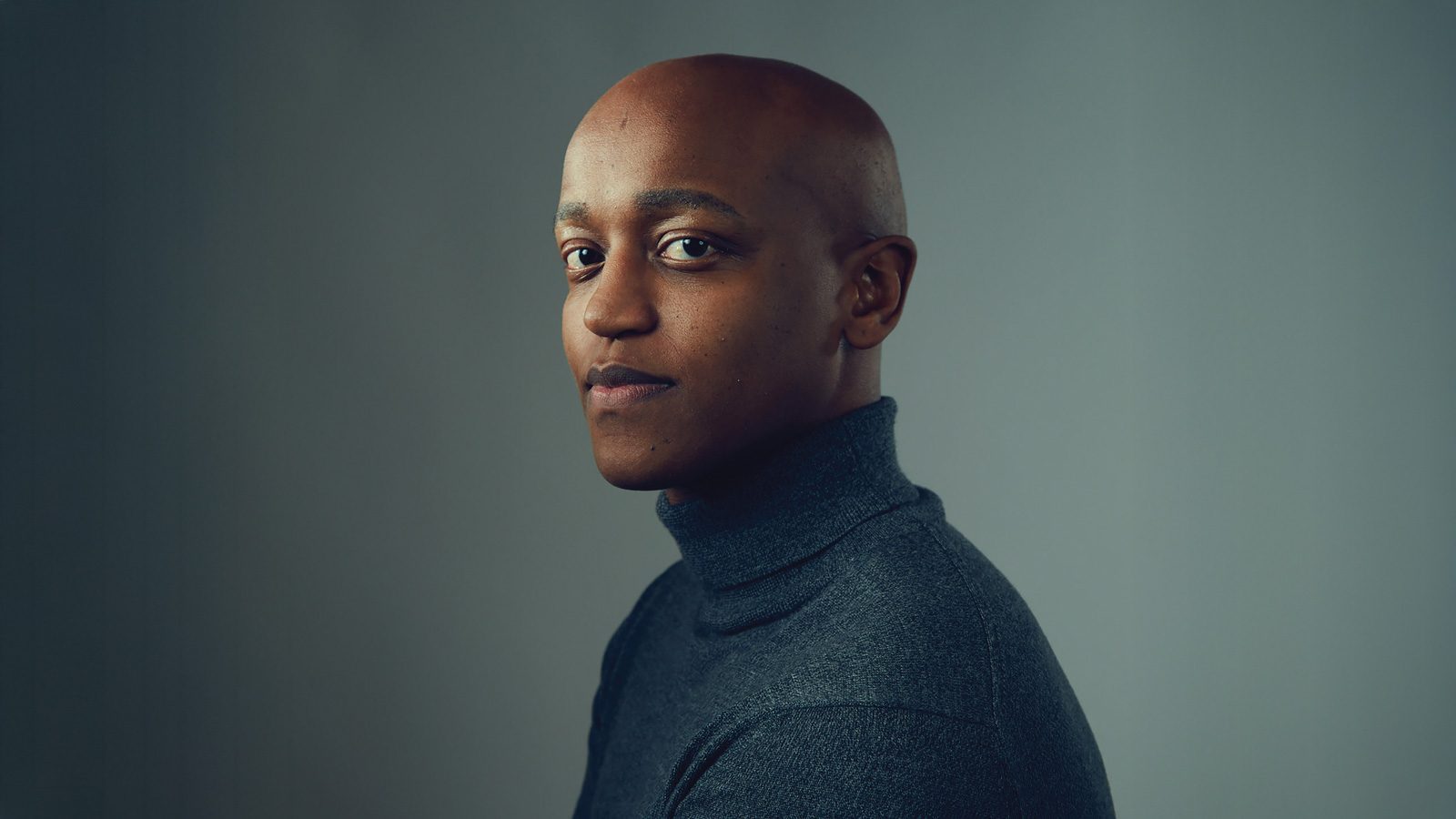 A person wearing a gray turtleneck, reminiscent of Girmay Zahilay's understated style, looks at the camera with a neutral expression against a plain background.