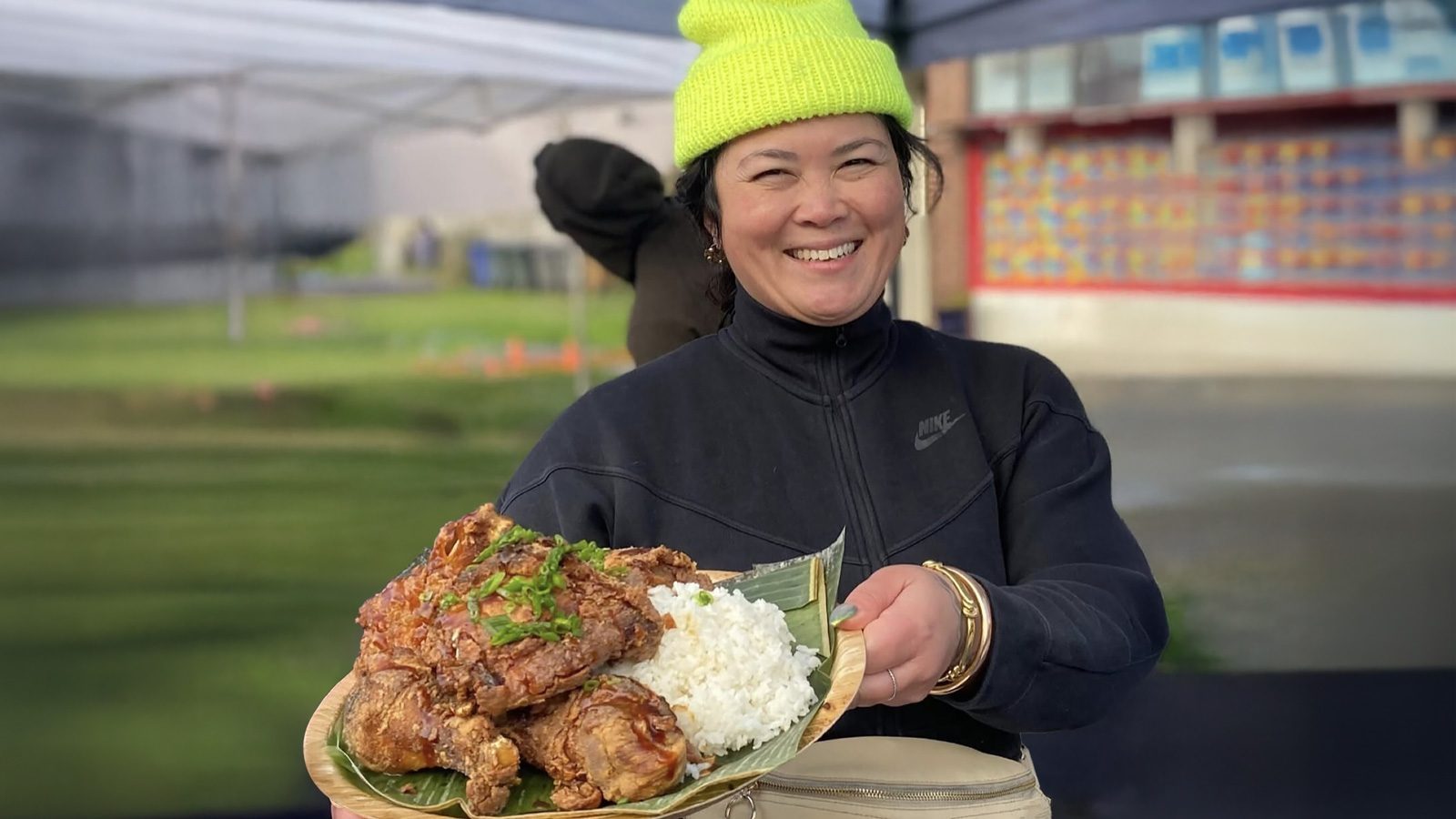 Under a canopy, Chef Jan Parker beams as they hold a plate of rice and cooked meat, artfully garnished with herbs. Dressed in a bright yellow beanie and dark jacket, they stand ready to share their culinary journey—a map of flavors from every corner they've explored.