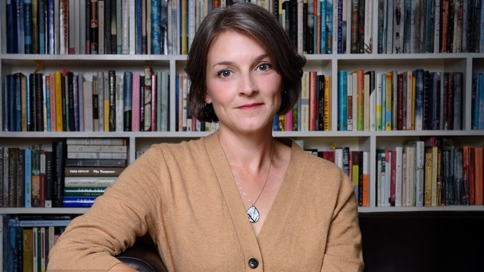 A person sits in front of a full bookshelf, wearing a tan sweater and a necklace, as if immersed in the secrets each book holds.