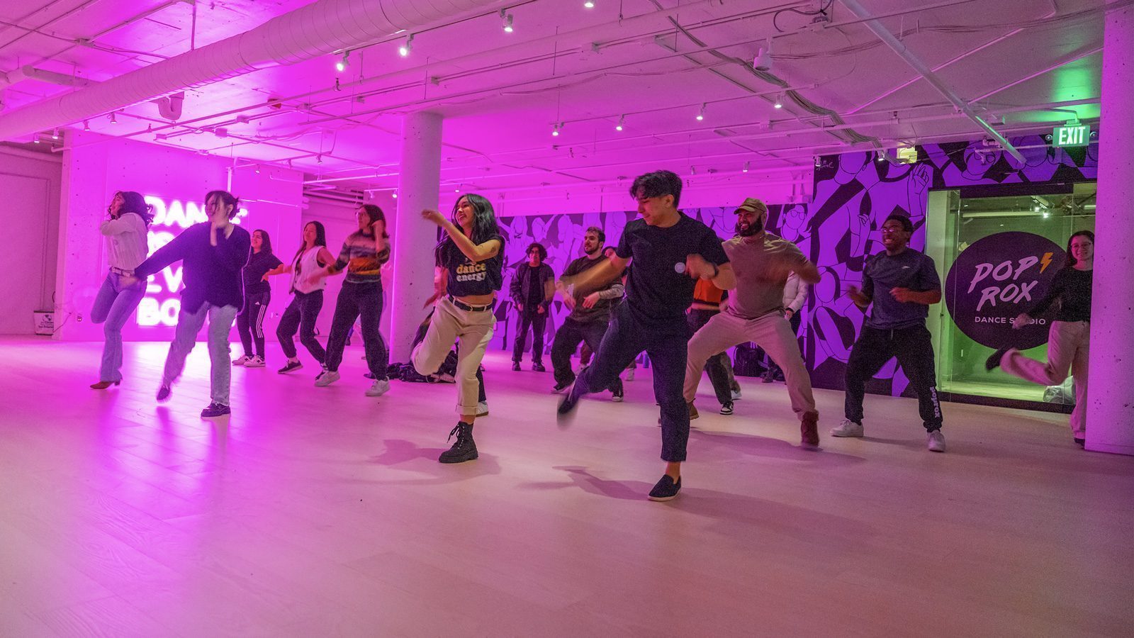 A group of people dancing in a studio with purple lighting and a neon sign in the background proudly displays their Courage to Move through every rhythm.