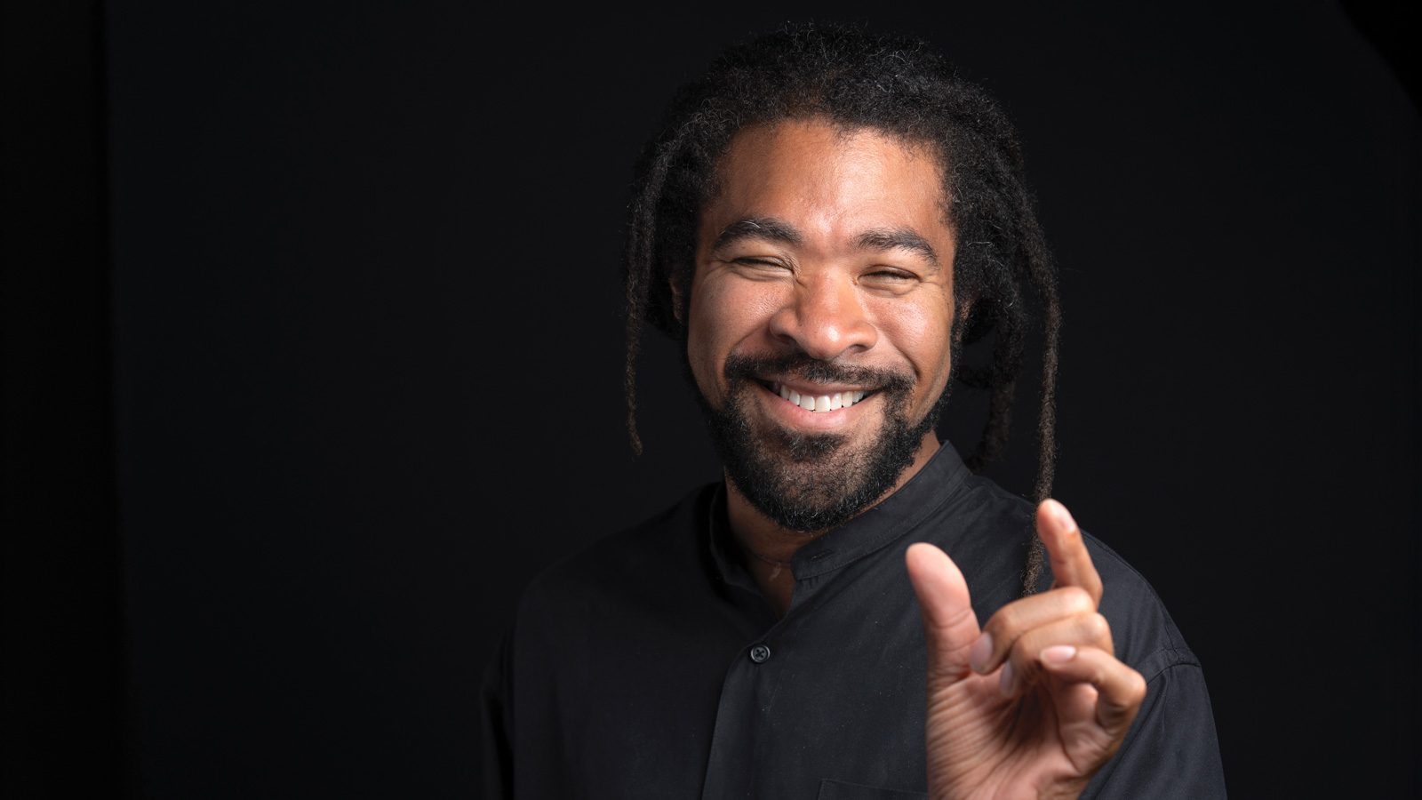 Rico Quirindongo, a smiling man with dreadlocks, holds his fingers apart in a black shirt against a dark background.