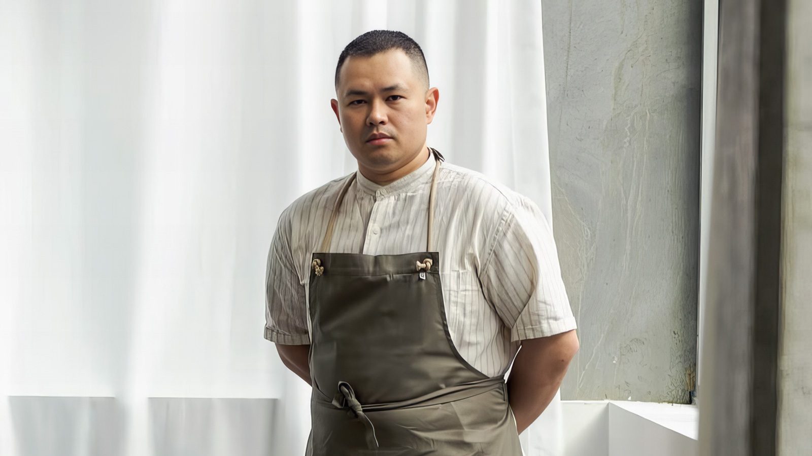 A person stands indoors wearing a white shirt and gray apron, embodying the essence of haute Cambodian food, as they look directly at the camera.