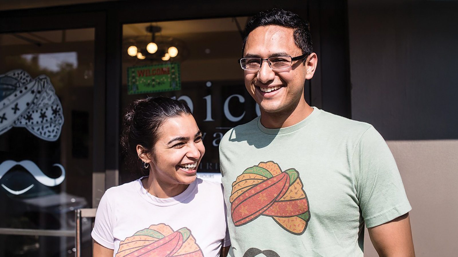 Two people smiling in front of a building entrance, both wearing shirts with taco graphics that echo the vibrant flavors Spice Waala brings with its authentic Indian street food to Seattle.