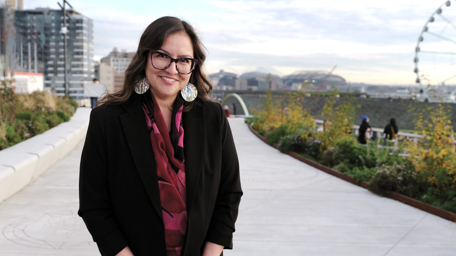Valery Segrest, wearing a black blazer and glasses, stands outdoors on a pathway with a cityscape and Ferris wheel in the background, passionately discussing native food systems.