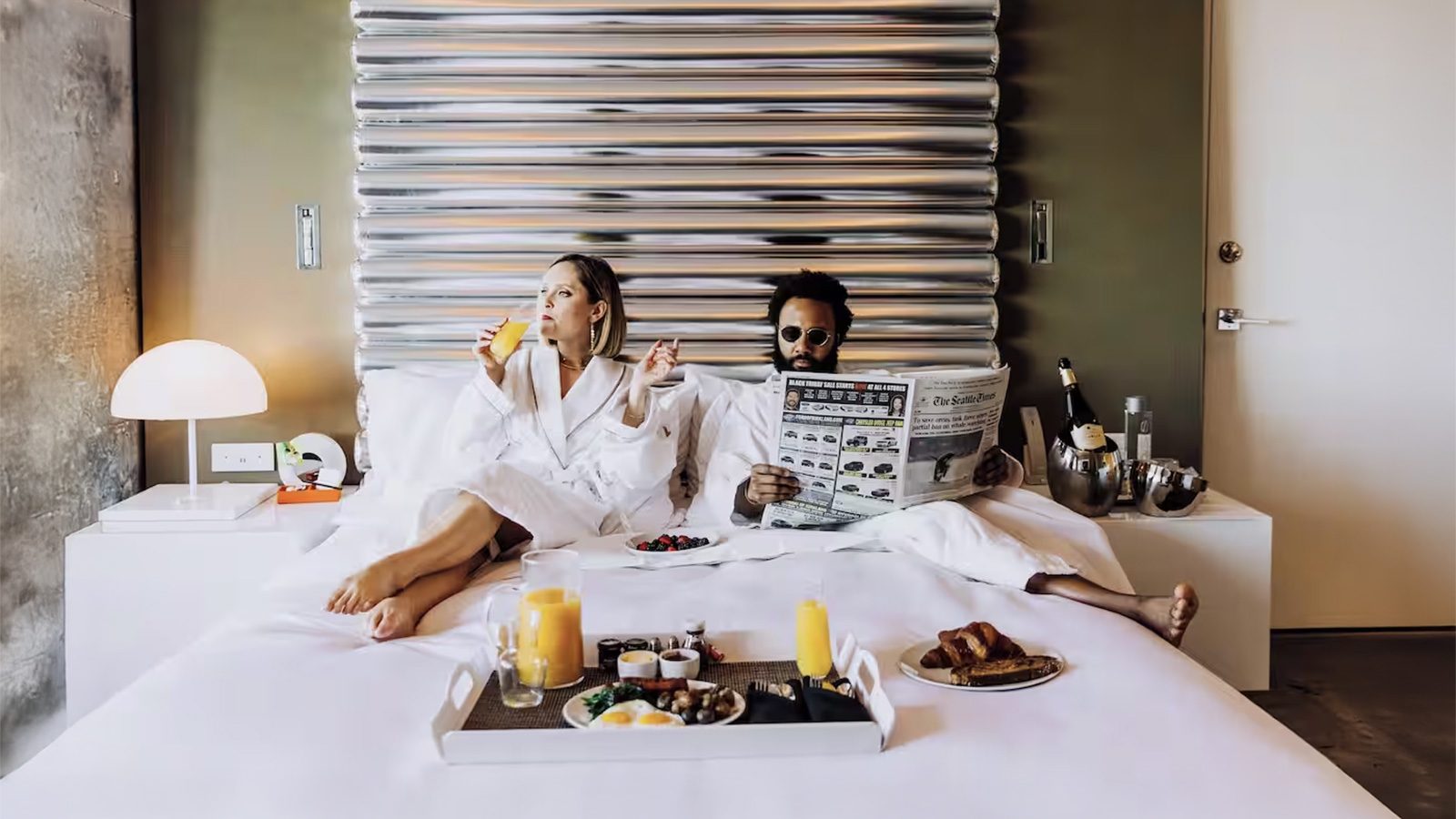 A couple in bathrobes relaxes in their luxurious suite, savoring a peaceful morning. One reads a newspaper while the other enjoys a glass of juice. A tray with breakfast treats adds to the perfect Valentine's Day atmosphere on the cozy bed.