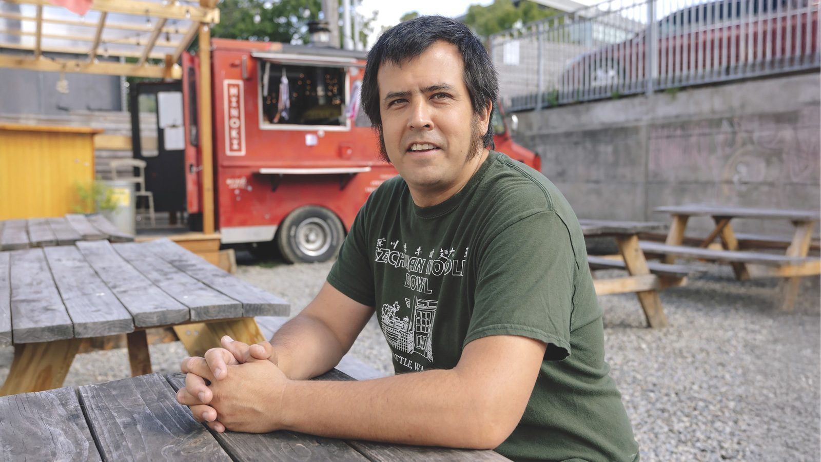 A person wearing a green T-shirt sits at a wooden picnic table in front of a red food truck, owned by restaurateur Yasuaki Saito who advocates for mental health.
