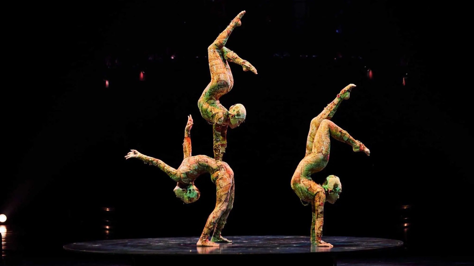 Three acrobats in colorful bodysuits perform a balancing act on a dark stage, each supporting one another in a dynamic pose, bending the limits of gravity with their grace and precision.