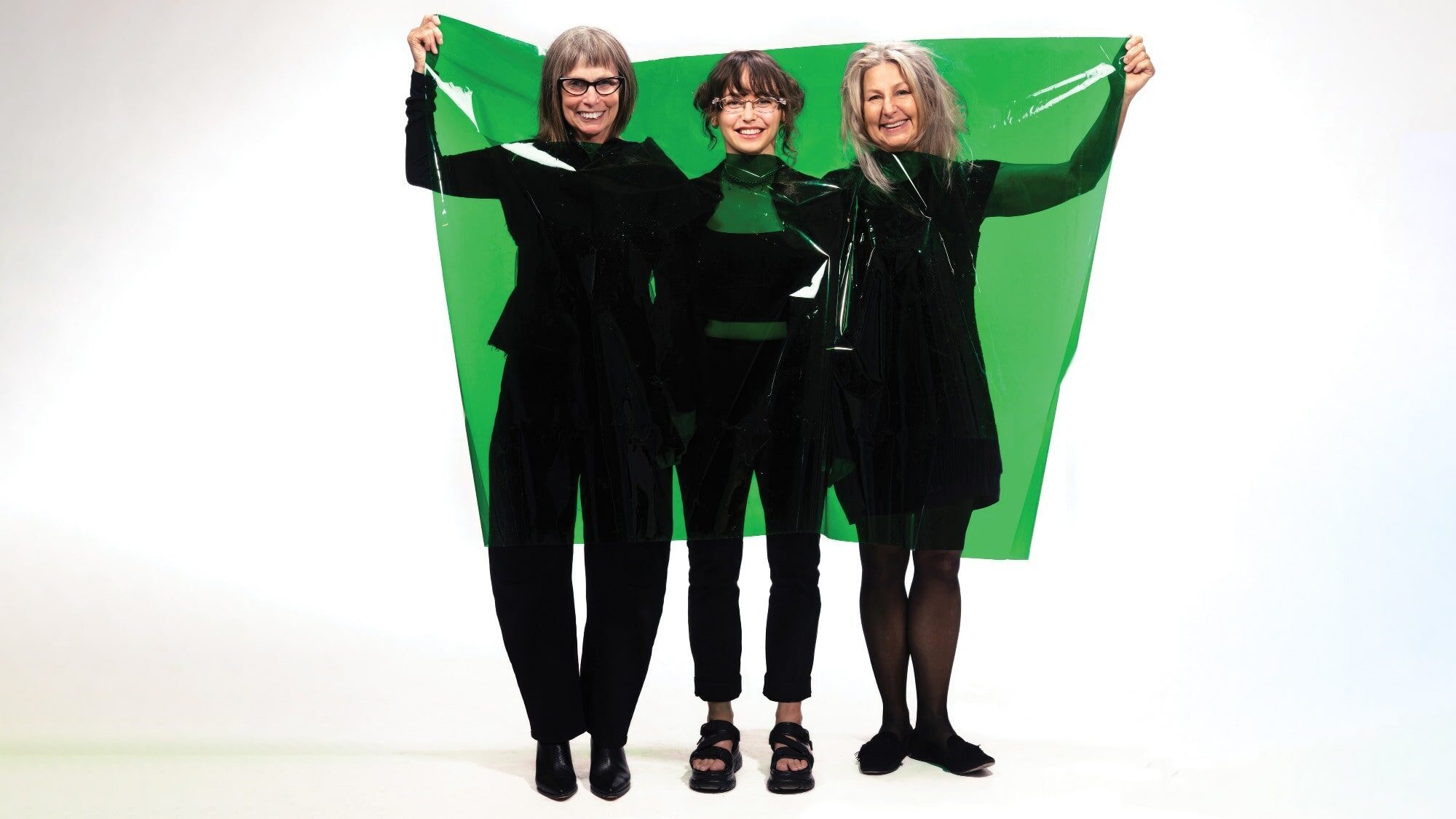 Three people stand together, embodying the spirit of Seattle creators, as they hold a large green transparent sheet in front of them against a plain white background. These philanthropists and art supporters unite to promote local artists boldly and passionately.