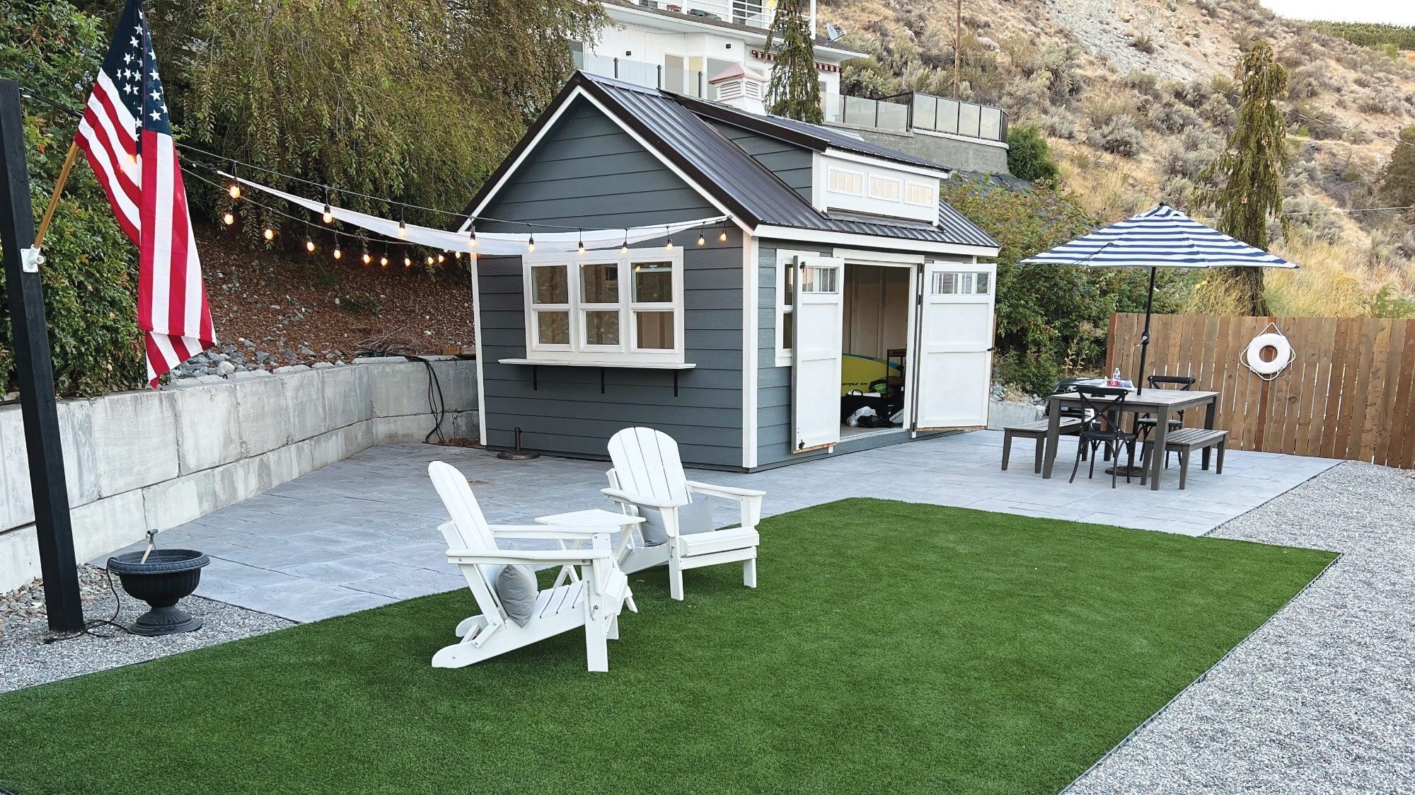 In a hilly backyard setting, this small grey shed with white trim features string lights and two white chairs on artificial grass. An outdoor table with a striped umbrella accompanies an American flag, capturing the charm of second homes in regions where Washington is a happy hunting ground for waterfront lots.