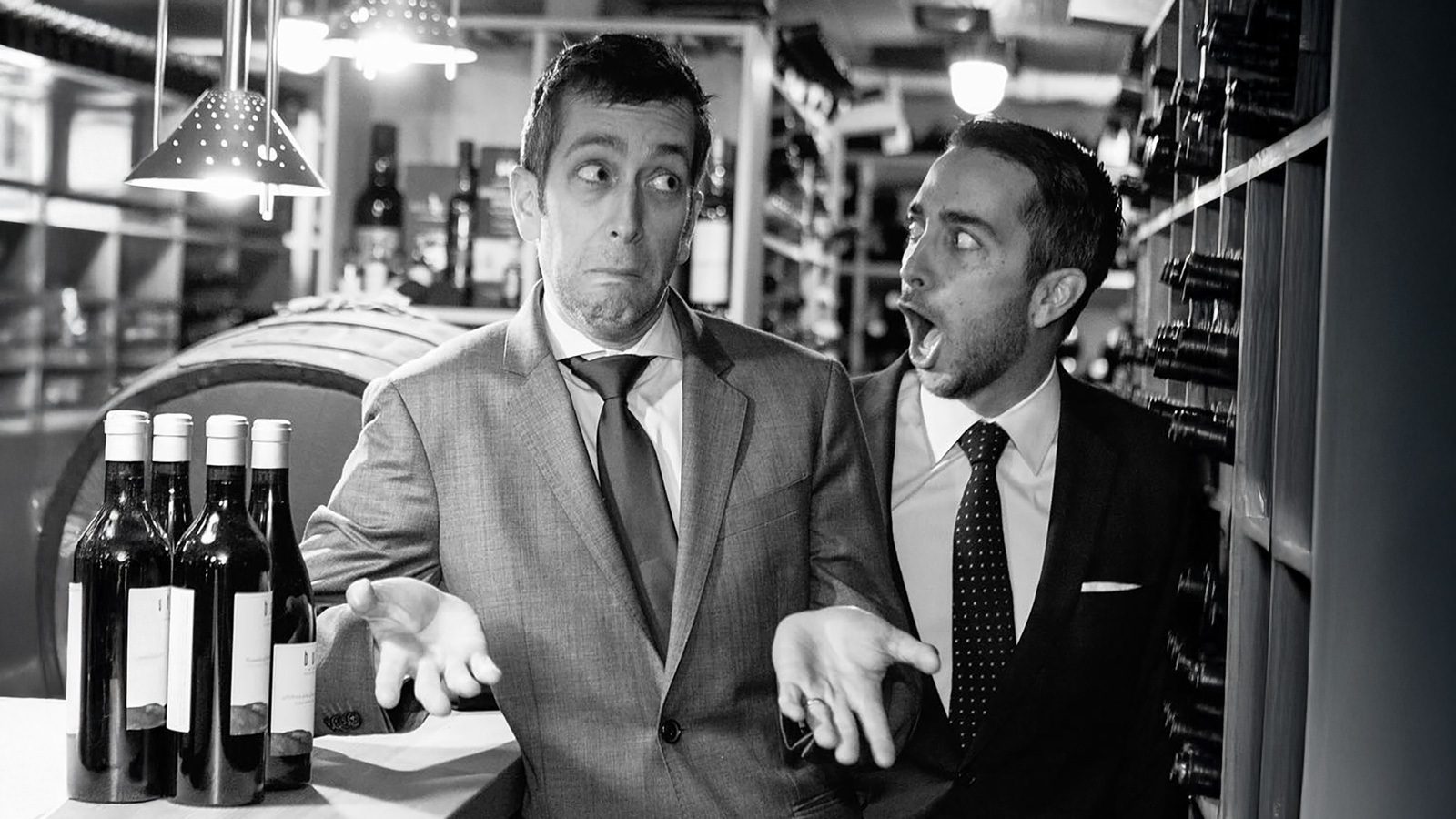 In the Canlis wine cellar, two men in suits stand perplexed amid bottles of wine, one shrugging in confusion while the other wears a surprised look, as if witnessing a culinary changing of the guard.