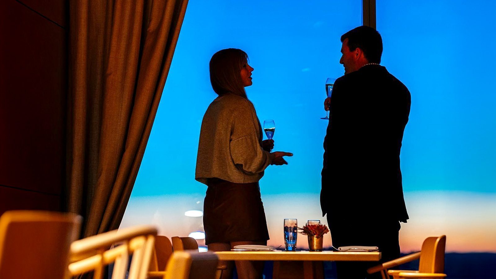 Two people stand by a window, holding glasses and chatting, with the vibrant colors of a Valentine’s Day sunset painting the scene in the background.