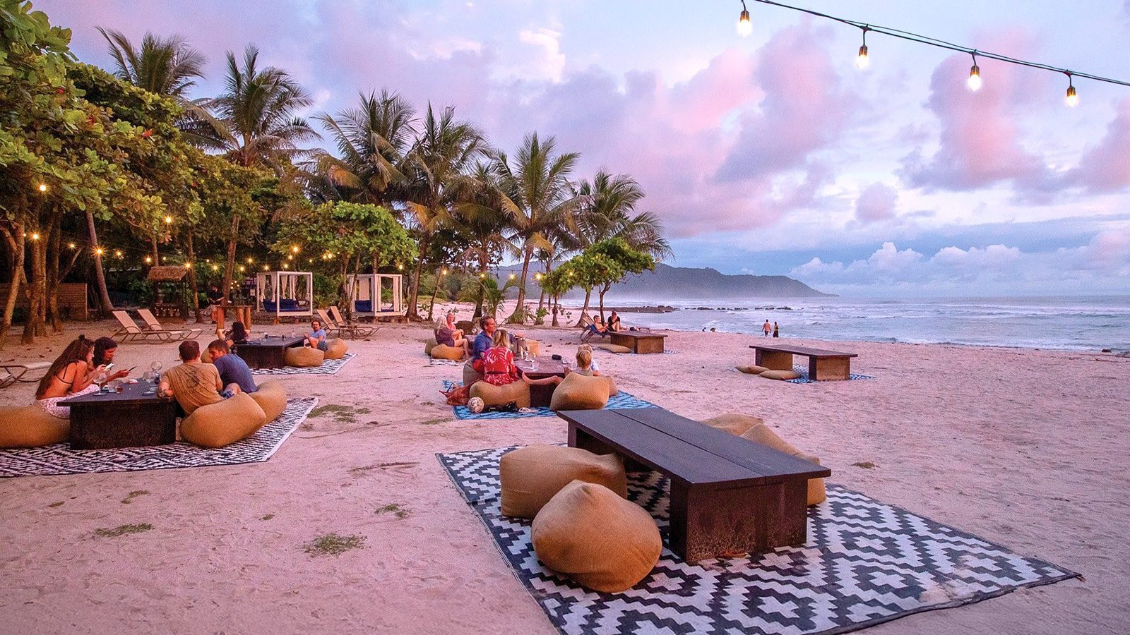 At sunset in Santa Teresa, Costa Rica, people unwind on bean bags and rugs beneath swaying palm trees and twinkling string lights, overlooking the serene ocean.