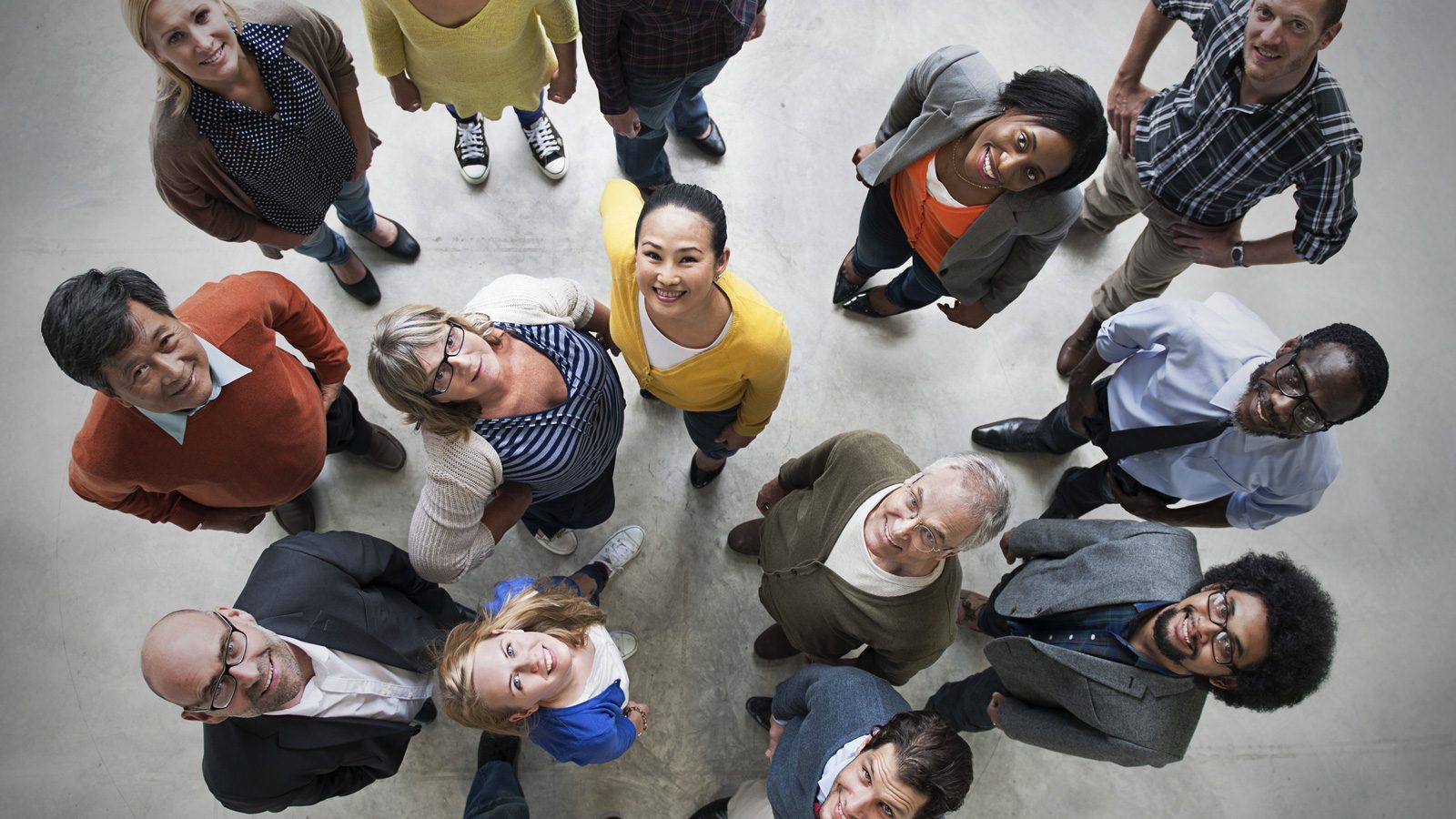 A diverse group of people, committed to DEI, stands together on a concrete floor, looking up at the camera, united in staying the course.