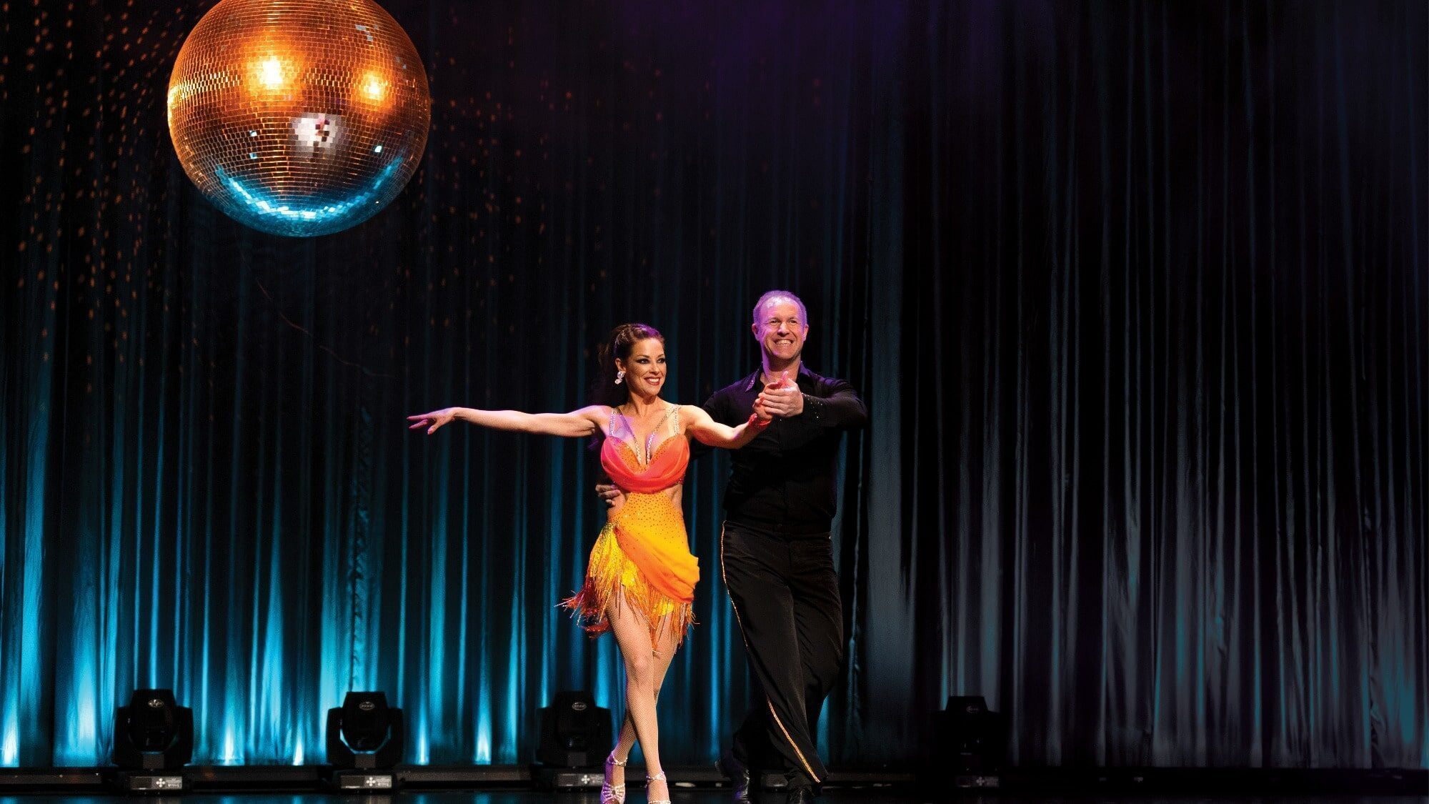 Under a large, illuminated disco ball, two dancers from Seattle Dances 2025 perform. The woman twirls in an orange and yellow dress while the man complements her in black attire. Their graceful movements aim to inspire change and support Plymouth Housing's mission to eradicate homelessness.