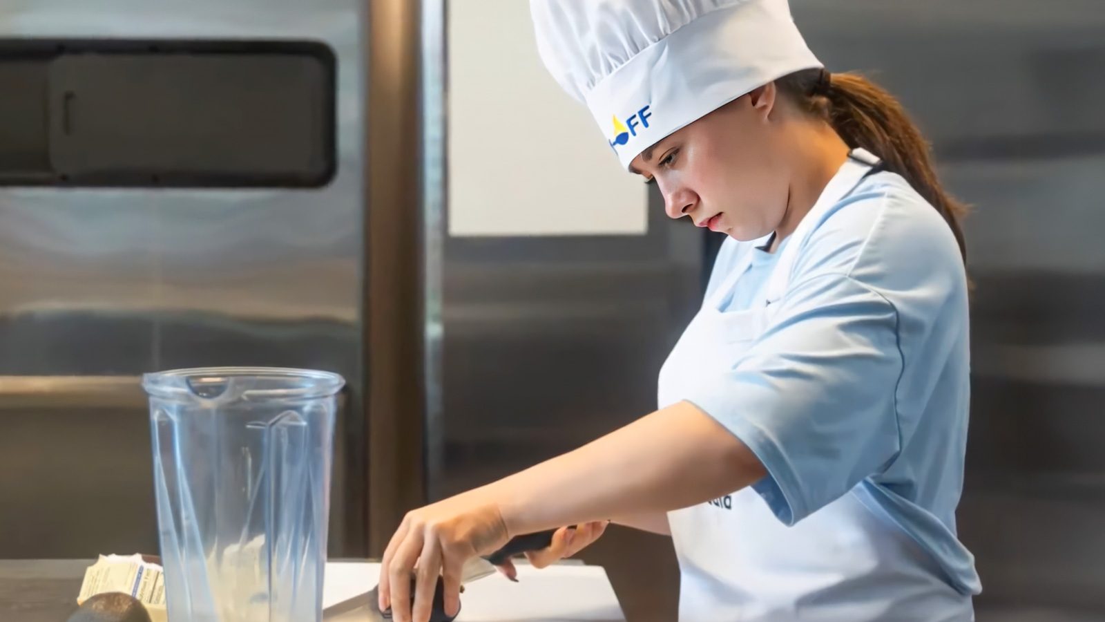 A 13-year-old girl in a blue chef uniform and hat is skillfully cutting an avocado beside a blender in the kitchen, fresh off her victory at a cooking competition.