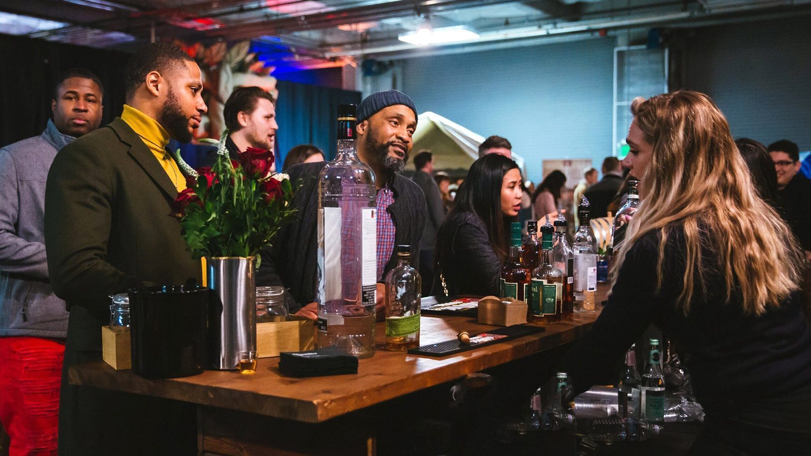 People standing at a bar counter, engaging in conversation with a bartender, surrounded by bottles and glasses during Seattle Cocktail Week.