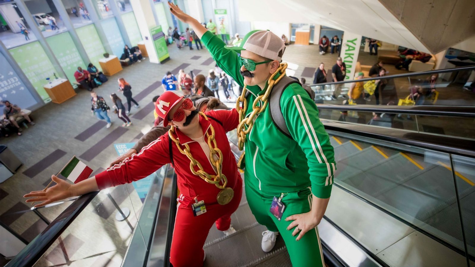 Two people in red and green tracksuits, with fake mustaches and oversized chain necklaces, pose on an escalator at a bustling event venue, capturing the eccentric spirit of Nerd Capital, America.