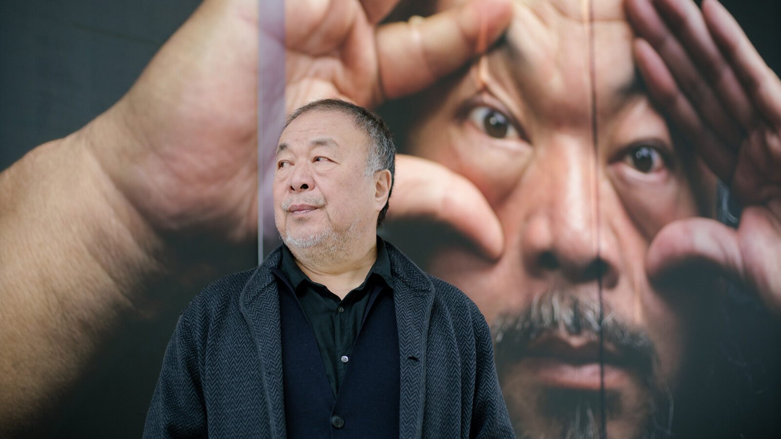 A man stands in front of a large artwork at SAM, part of Ai Weiwei's biggest U.S. show, depicting a close-up of another man's face with hands framing his eyes.