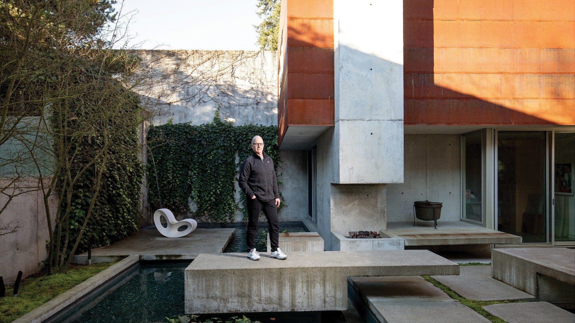 A person stands on a concrete platform in a modern architectural courtyard, designed by architect Eric Cobb, featuring a pool, vine-covered walls, and a distinctive chair. This serene space exemplifies the allure of a meticulously crafted second home.