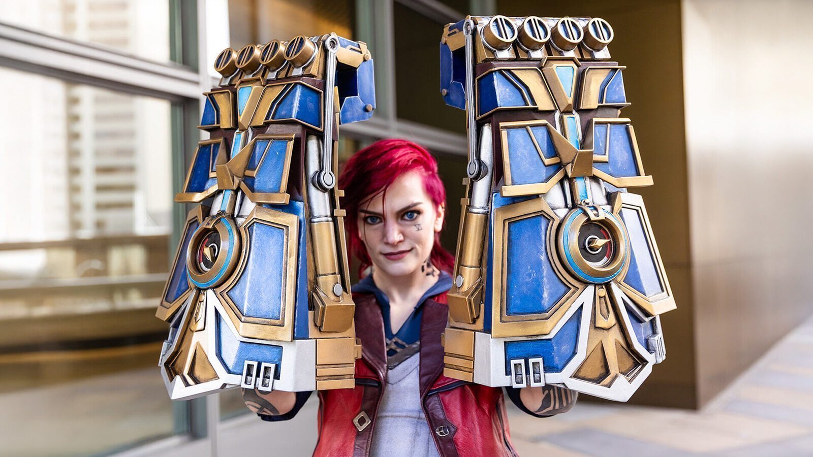 Person with red hair wearing large, intricately designed blue and gold gauntlets, standing in front of a building with glass windows.