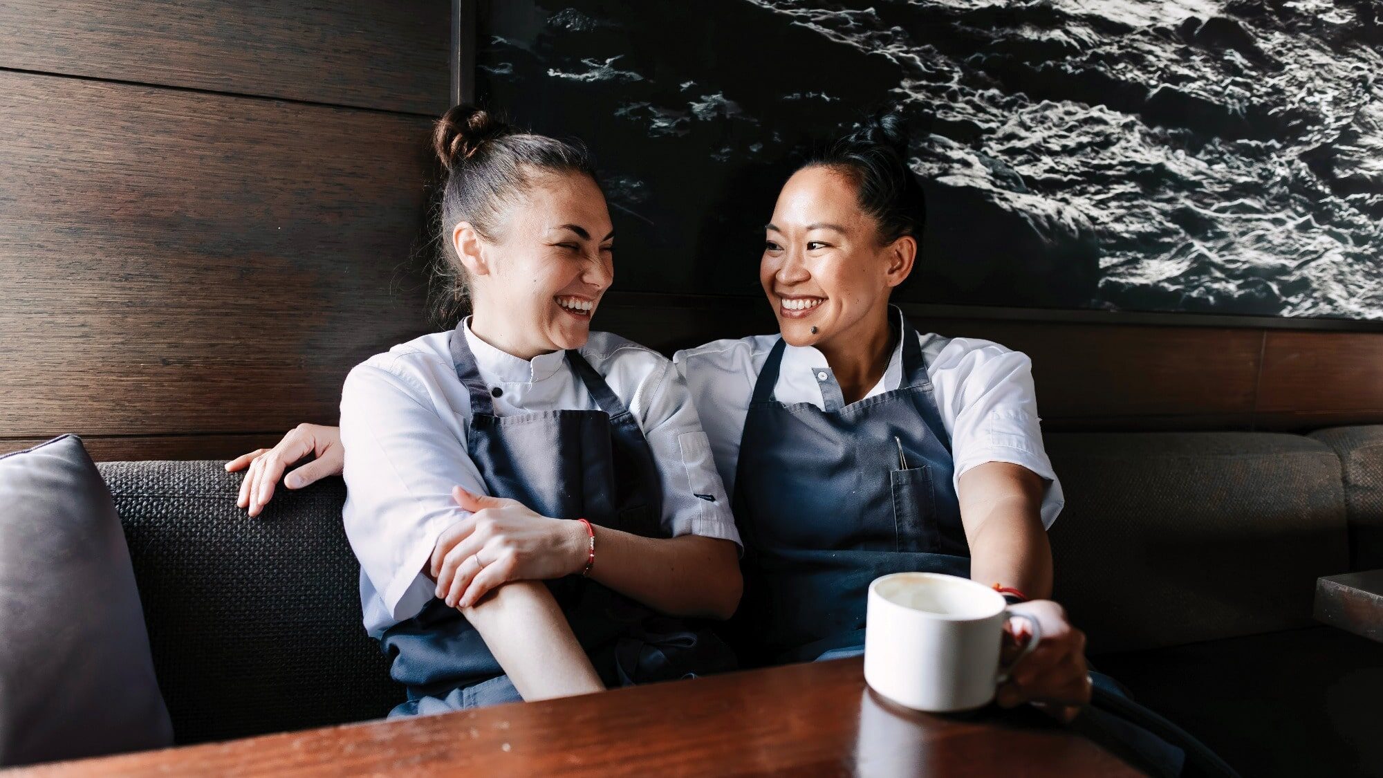 Two people in chef uniforms sit on a couch, smiling and looking at each other as if sharing a secret recipe. One holds a mug, their chemistry palpable. They are in a relaxed setting with a dark textured wall in the background, adding to the romantic ambiance.
